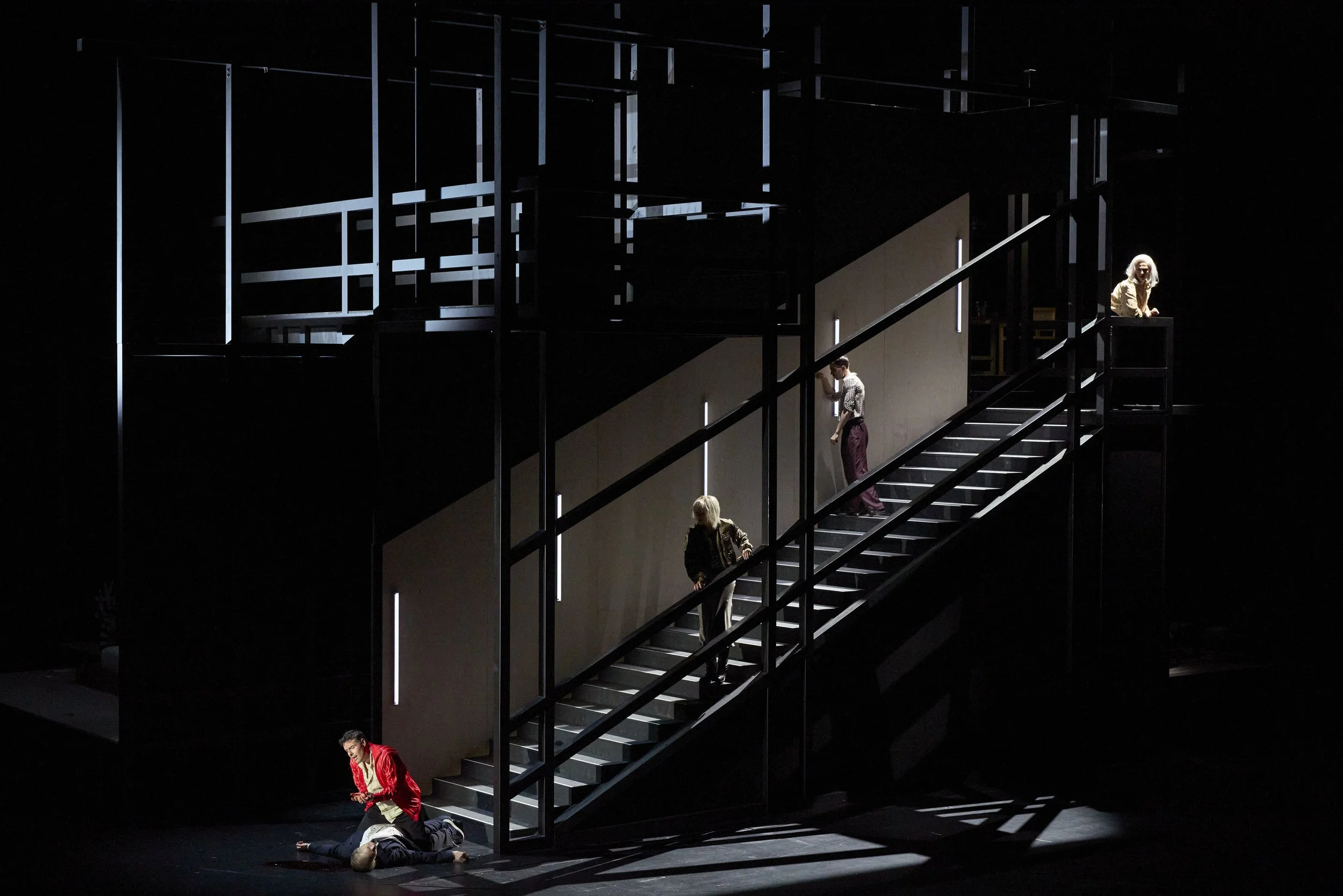 Theaterbühne mit schrägem schwarzem Treppenhaus, auf dem vier Schauspieler in verschiedenen Positionen stehen, einer liegt am Boden, während andere auf der Treppe und einer auf einem Balkon stehen.