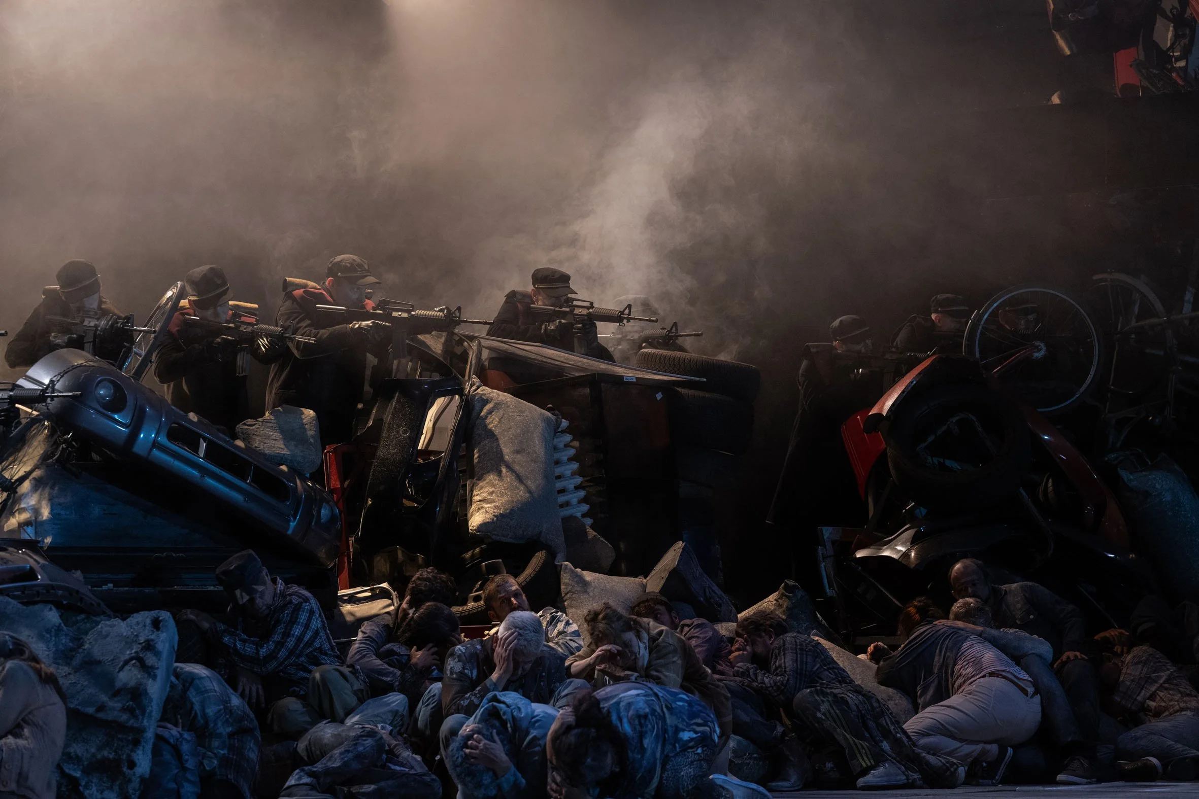 Menschen, die im Konflikt auf dem Boden liegen, während bewaffnete Streitkräfte mit Waffen in der Hand in den Hintergrund schauen, bei einer Szene mit Rauch und Trümmern.