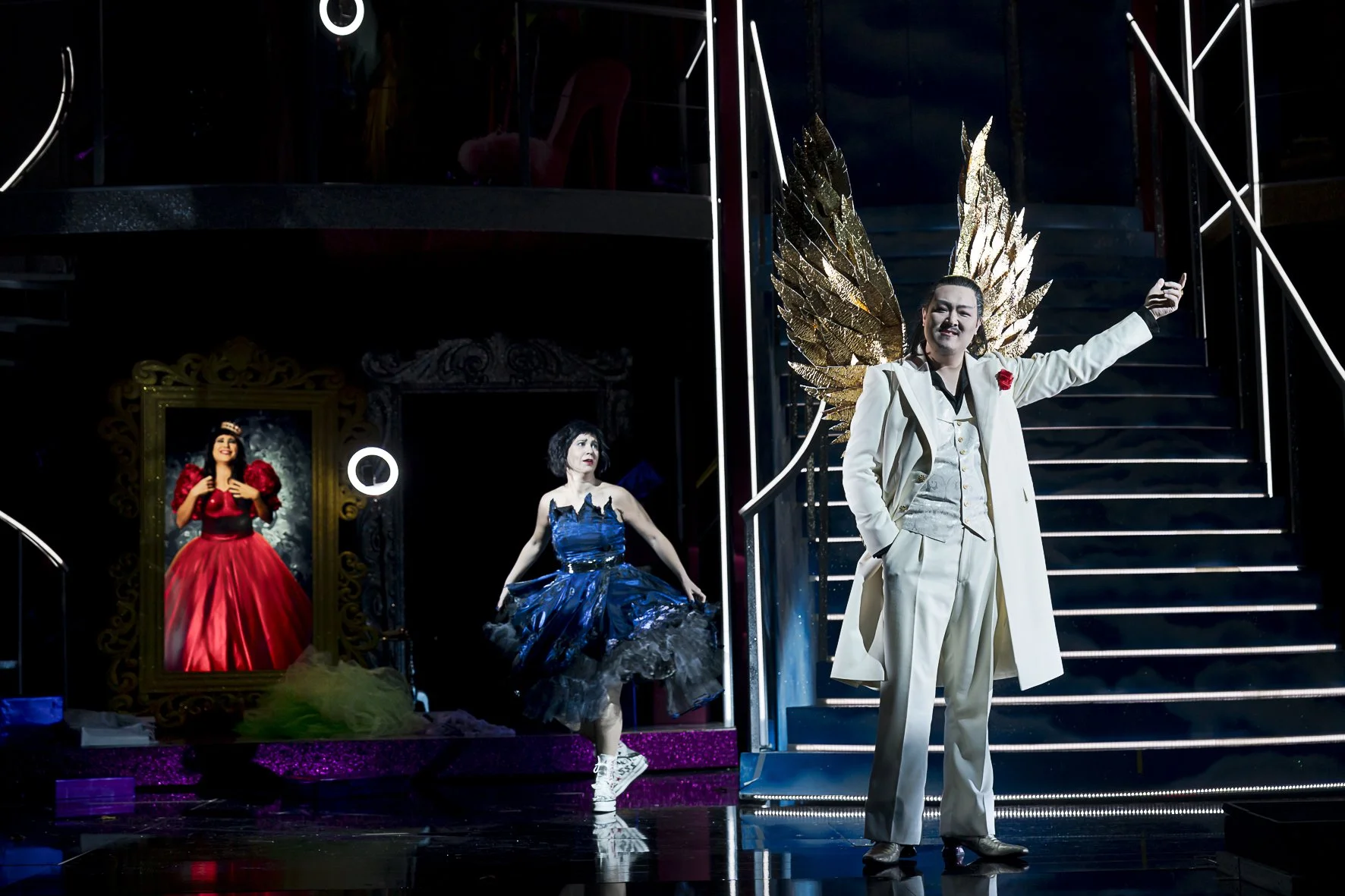 Drei Personen in Kostümen auf einer Bühne: eine Person in einem weißen Anzug mit großen goldenen Flügeln, eine Person in einem grünen Tutu, und eine Person in einem roten Ballkleid, vor einem dunklen Hintergrund mit Spiegel und Stufen.