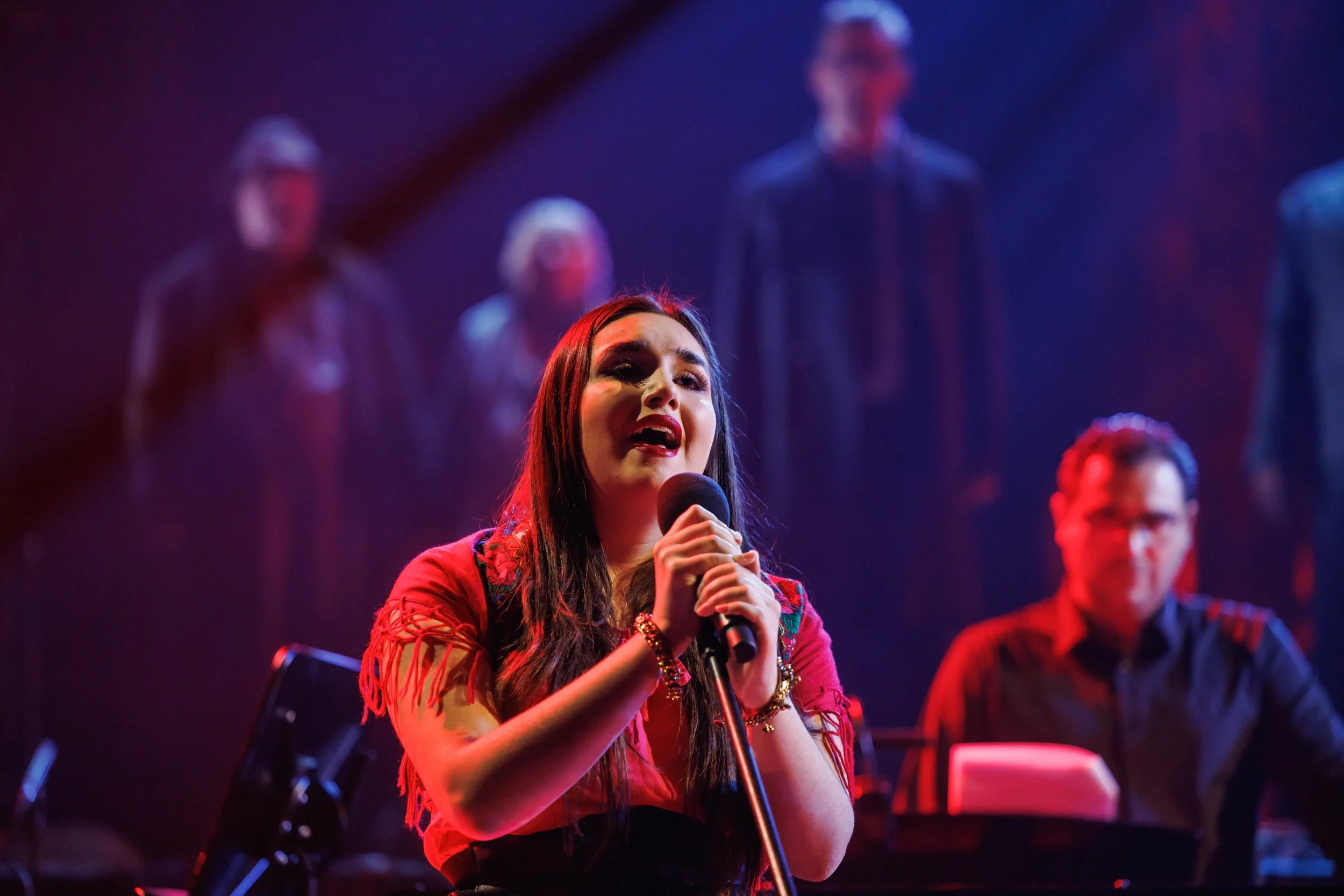 Eine Frau singt mit einem Mikrofon auf einer Bühne, im Hintergrund sind fünf Musiker in dunkler Kleidung zu sehen, unter farbigem Bühnenlicht.