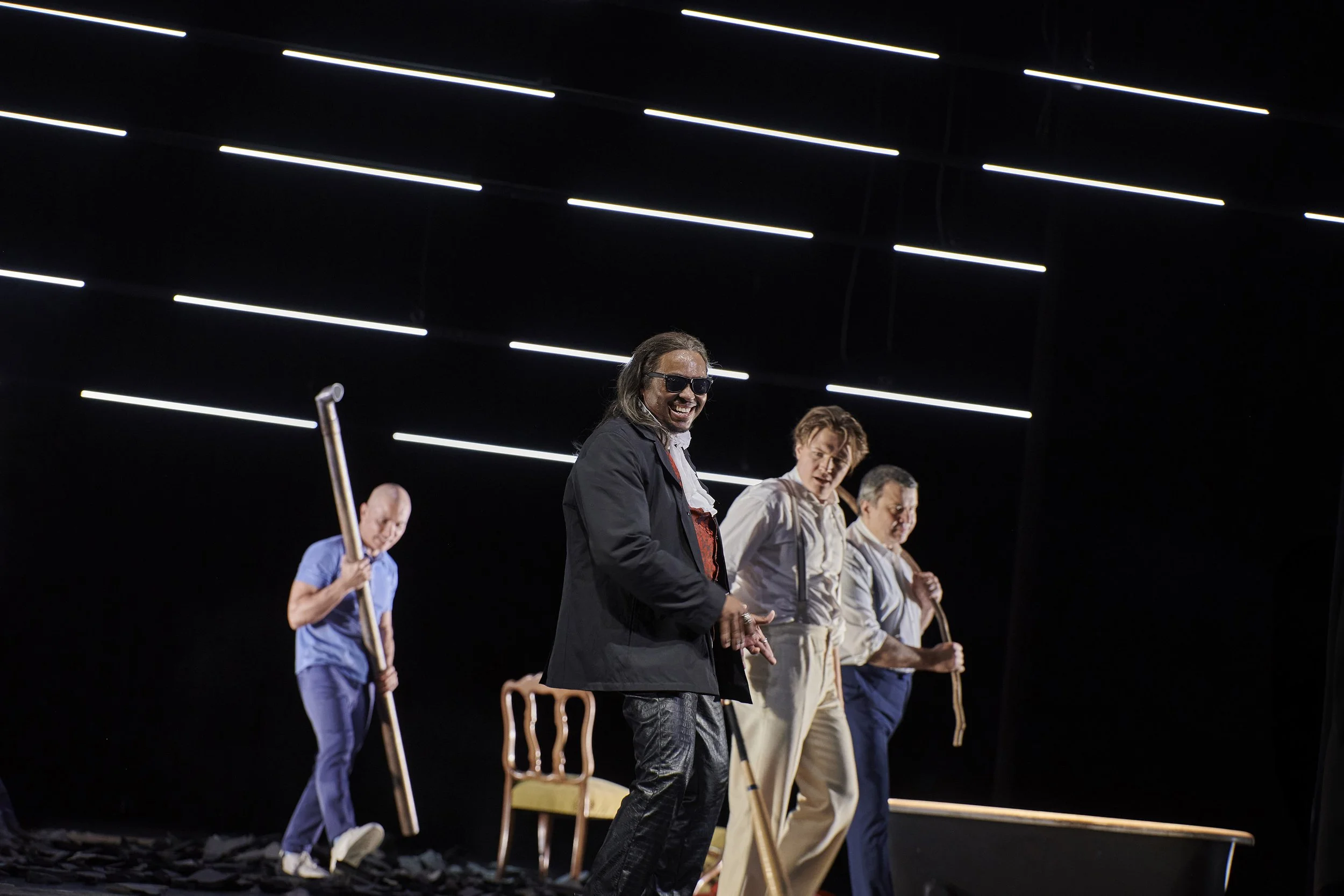 Vier Schauspieler auf einer Bühne, wobei einer im Vordergrund lächelnd schaut und die anderen drei im Hintergrund mit Stöcken stehen, bei dunklem Hintergrund mit horizontalen Lichtlinien.