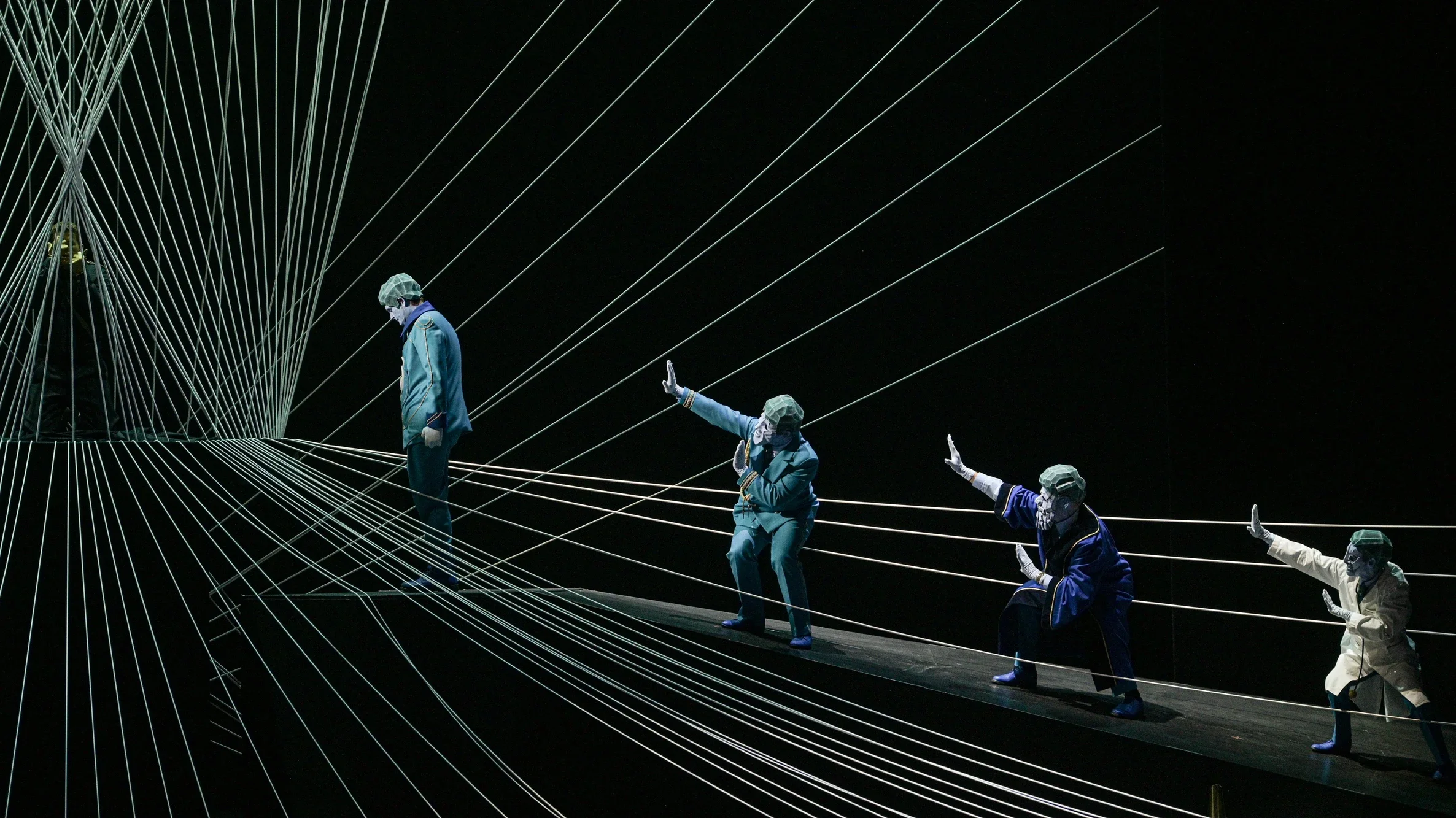 Bühnenaufführung mit fünf Darstellern, die auf einem schmalen Steg mit weißen Fäden im Hintergrund agieren, alle tragen Schutzmasken und historische Kostüme.