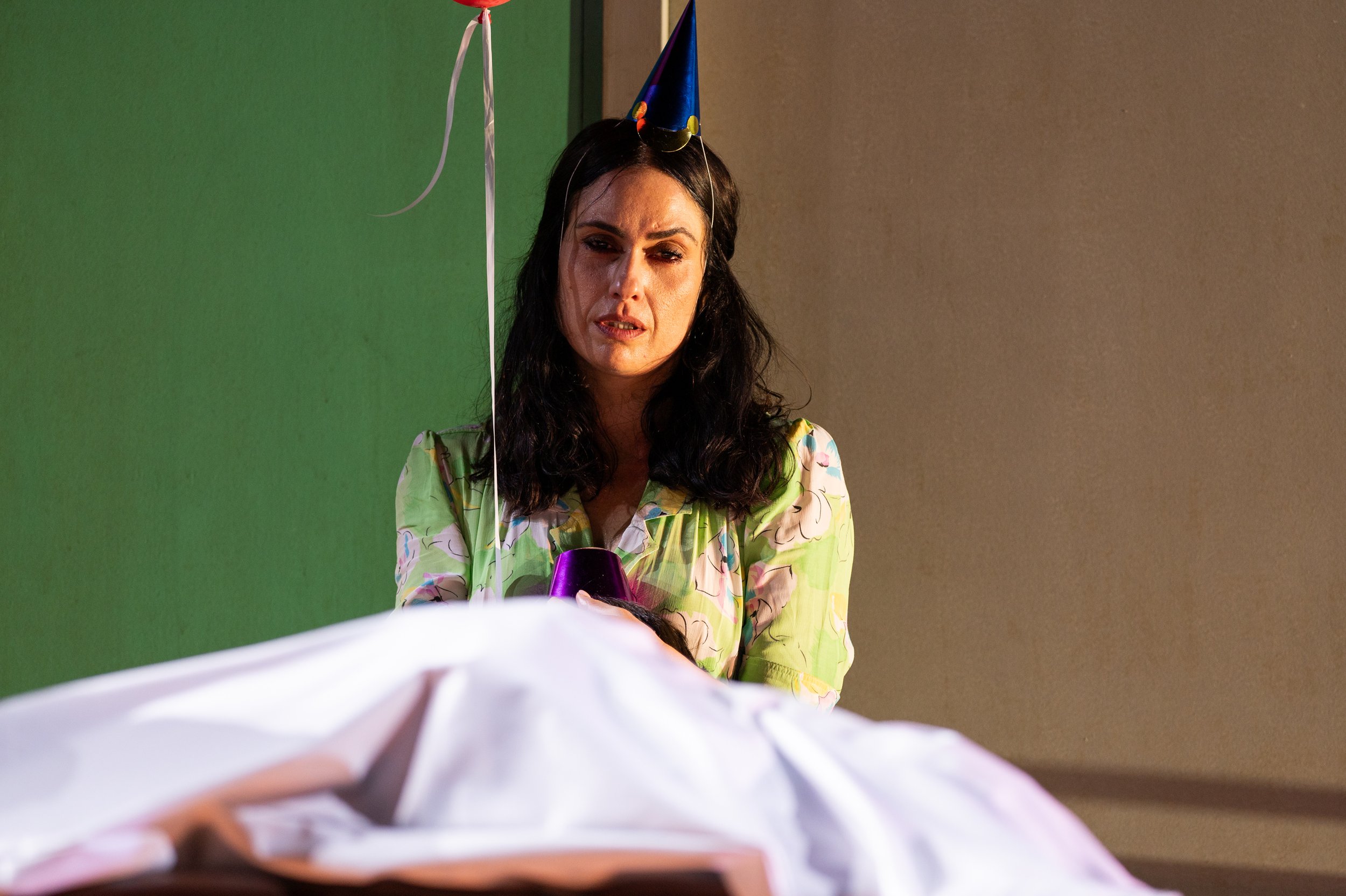 Frau trägt eine Partyhütchen, schaut traurig auf ein Begräbnis. Es gibt eine offene Sarg, darüber ein weißes Tuch, im Vordergrund.