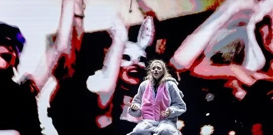 Eine junge Frau, die auf einer Bühne sitzt, umgeben von einem großen Bildschirm mit einem Hund und einer Person, die eine Maske trägt.