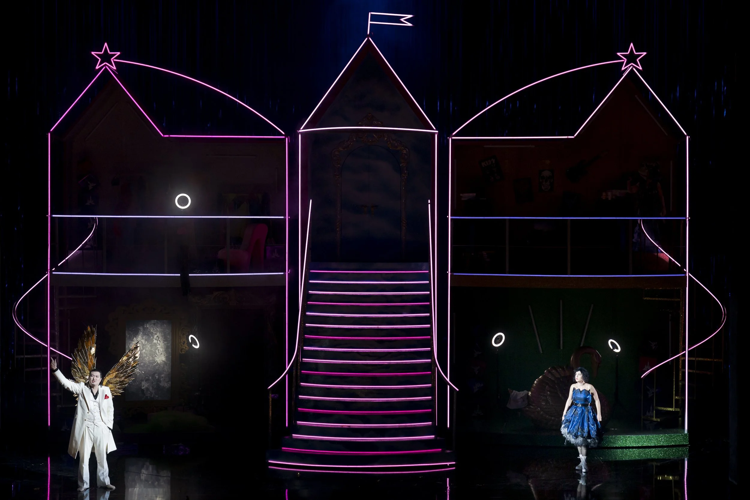 Bühne mit Neon-Kulisse, zwei Personen im Kostüm, eine mit Flügeln, die andere in blauer Robe, bei einer Theateraufführung.