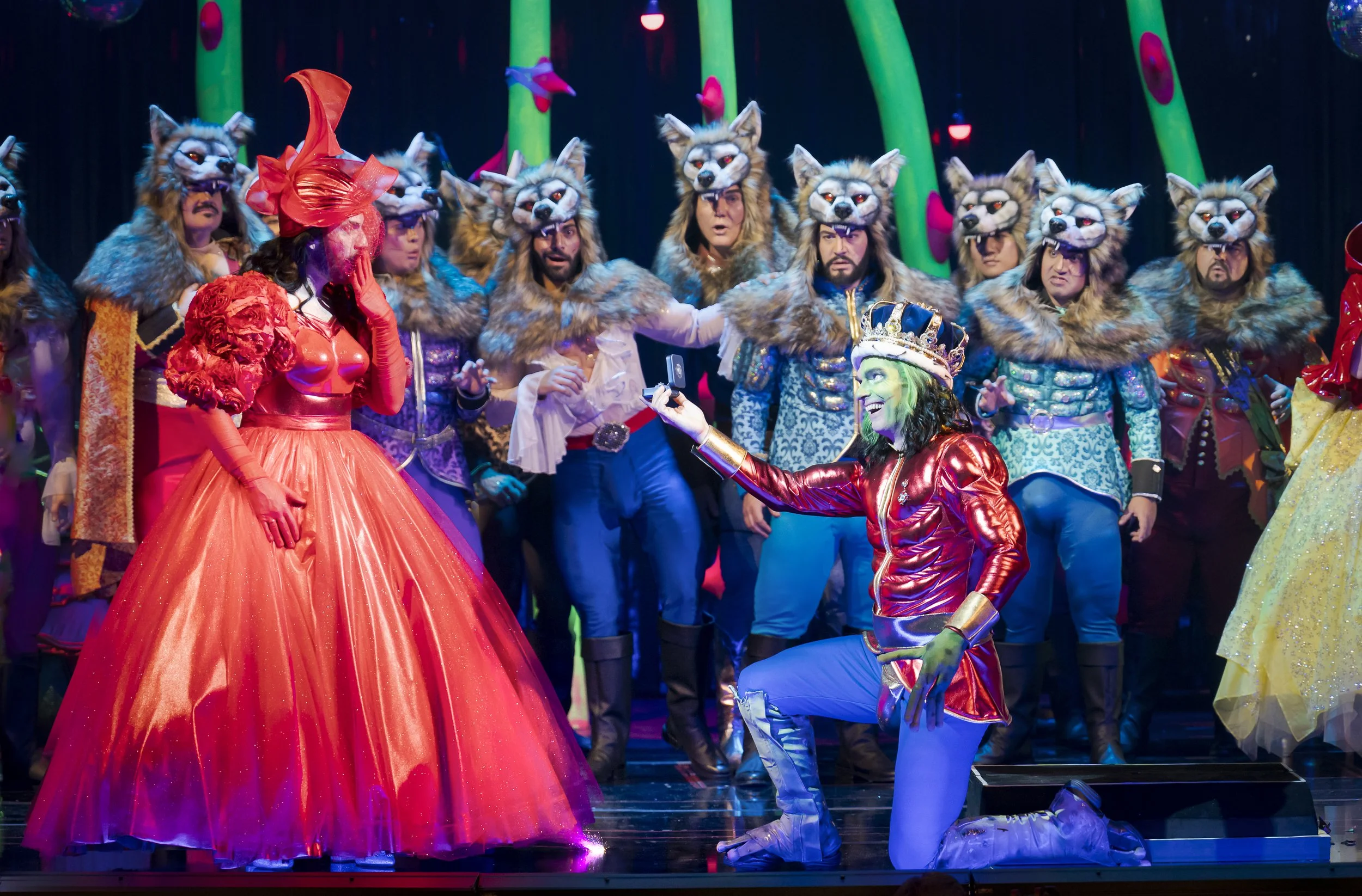Menschen in Tierkostümen und eleganter Kleidung auf einer Bühne, bei einer Theatervorstellung oder einem Karneval, mit bunten Kulissen im Hintergrund.