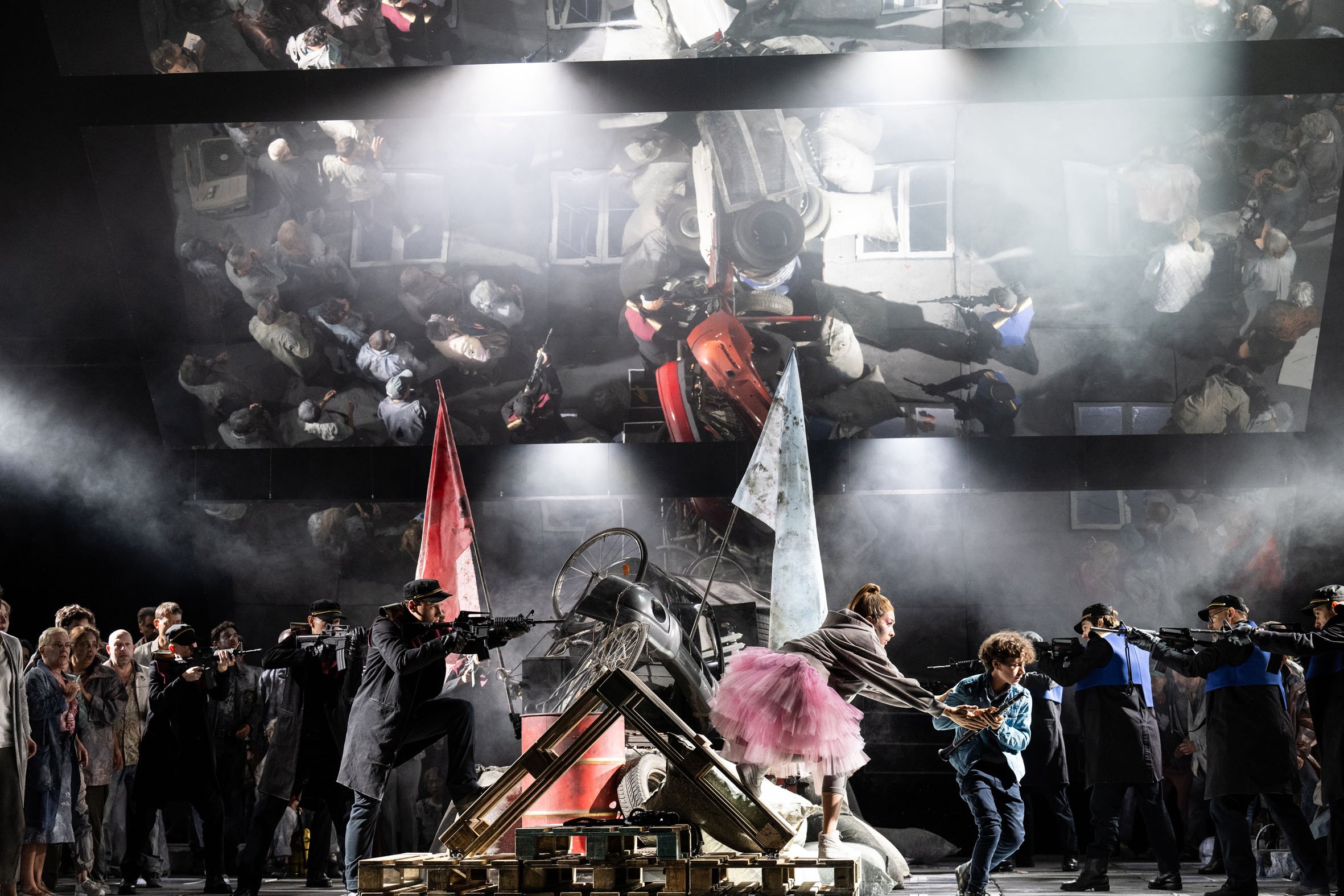 Theateraufführung mit einer Szene voller Chaos, Menschen, Waffen, Flaggen, Rauch und einer großen Spiegelwand. Schauspieler in Anzügen und Kostümen, maskierte Personen, Requisiten und eine komplexe Bühnenkulisse.