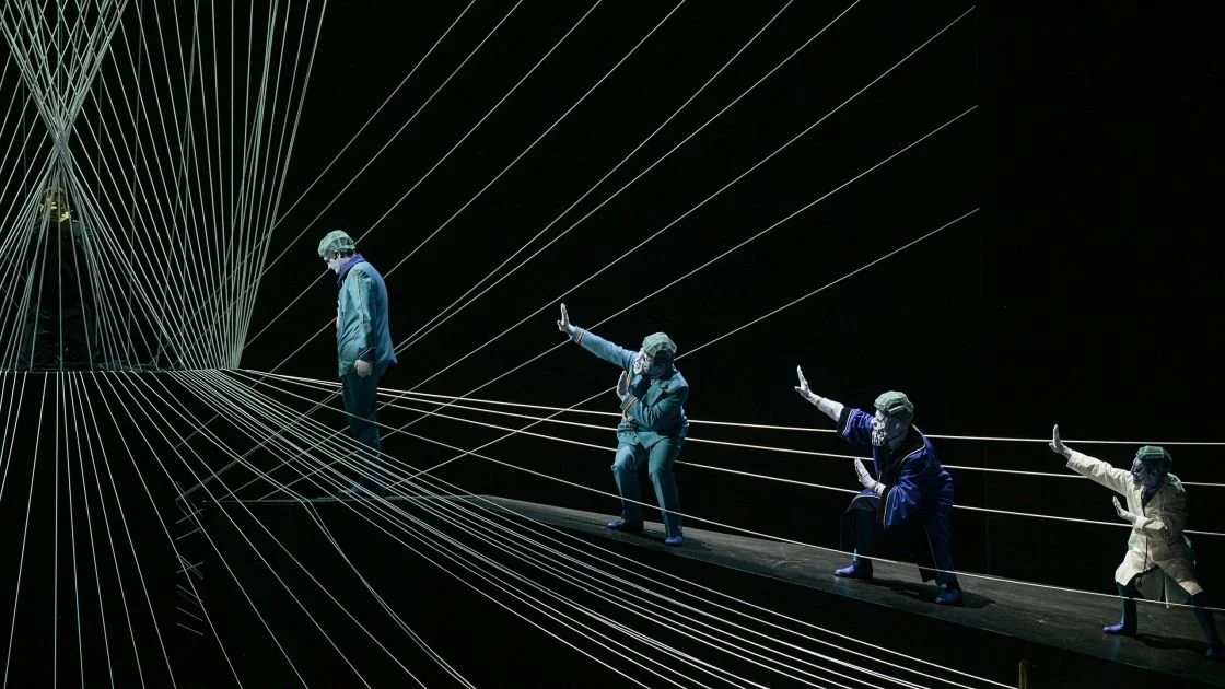 Bühnenperformance mit fünf Personen in Schutzkleidung auf einem schmalen Steg, umgeben von weißen Linien, die ein Netz bilden, gegen einen schwarzen Hintergrund.