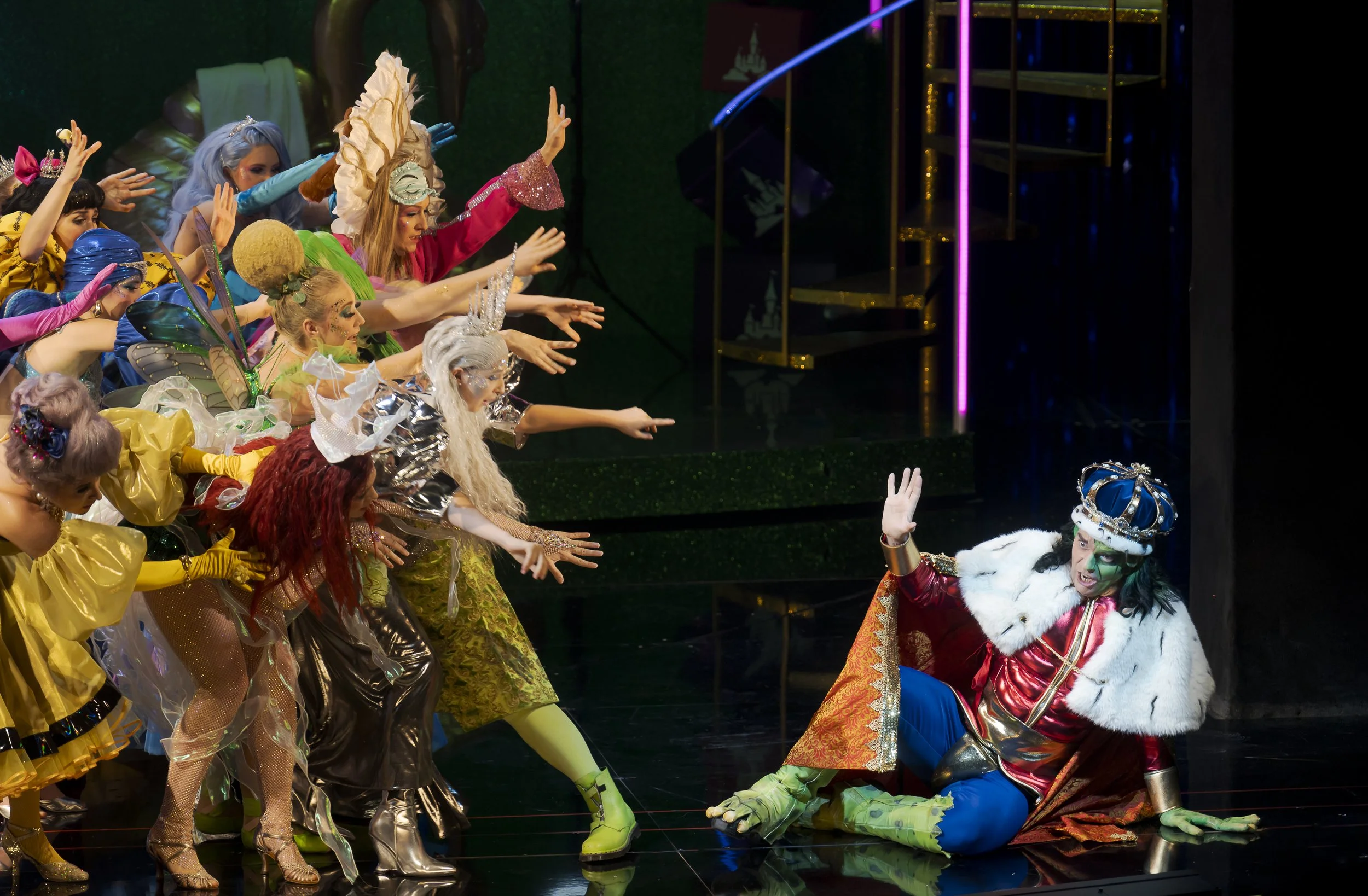 Schauspieler in bunten, fantasievollen Kostümen auf einer Bühne, eine Person in einer Königs-Kostüm auf dem Boden sitzend, die andere Person zeigt mit der Hand eine Geste der Ablehnung oder Warnung.