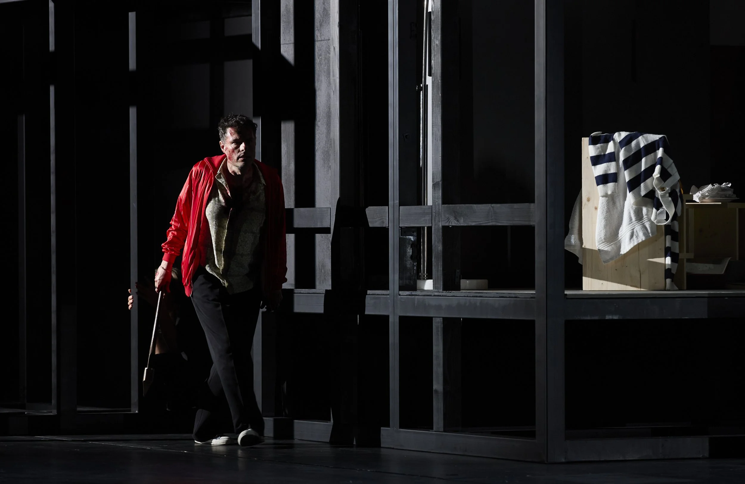 Eine Person mit dunklen Haaren, weißem Hemd, roter Jacke, schwarz Hosen, vor einer dunklen Kulisse, die eine schwarze Wand mit geometrischen Mustern zeigt, trägt eine Brille und hält einen Gehstock.