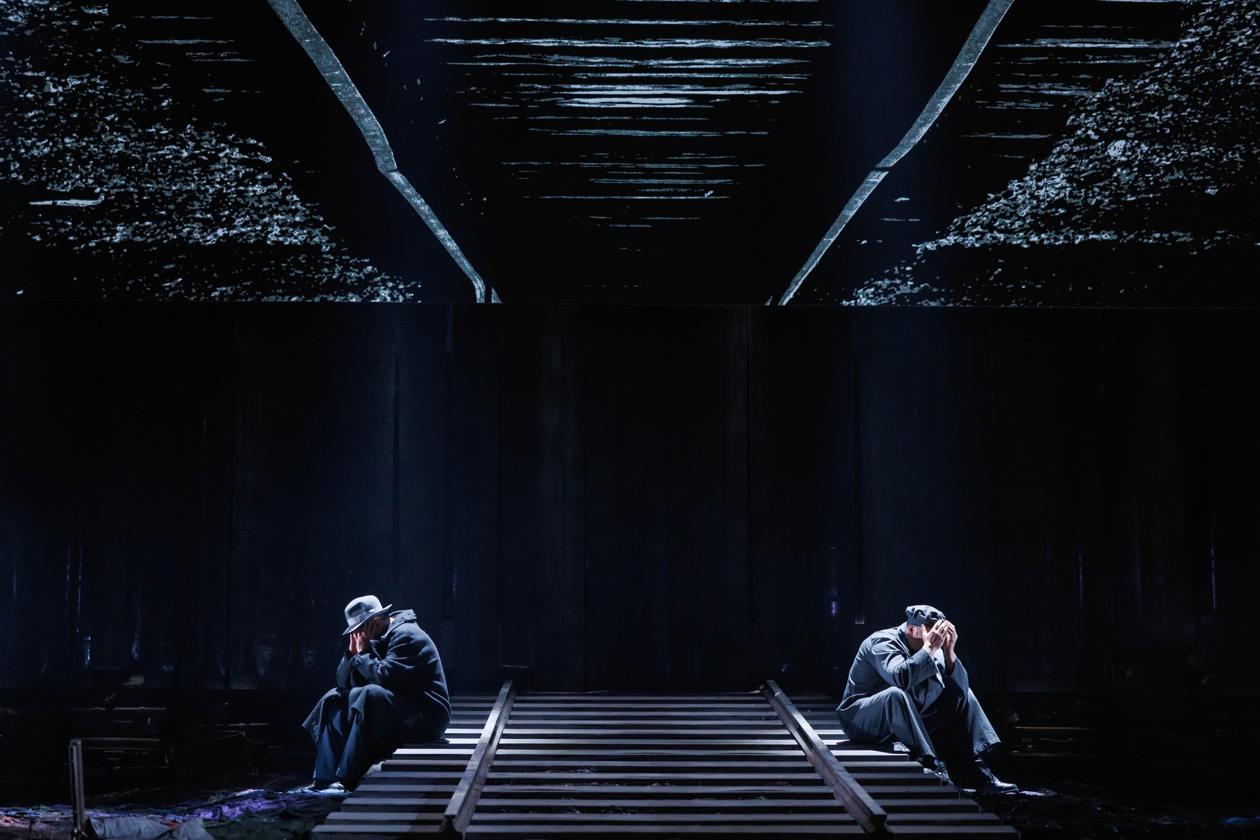 Zwei Männer sitzen auf Holzbahnen, in einer dunklen, minimalistischen Umgebung, jeder mit den Händen am Kopf, im Hintergrund eine reflektierende Deckenfläche.