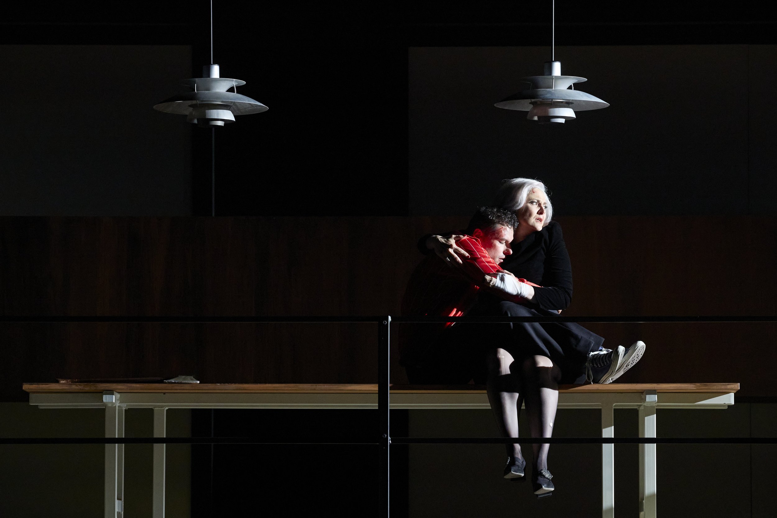 Zwei Menschen sitzen auf einer Bank, umarmt in einer dunklen Umgebung, beleuchtet von zwei Deckenlampen.