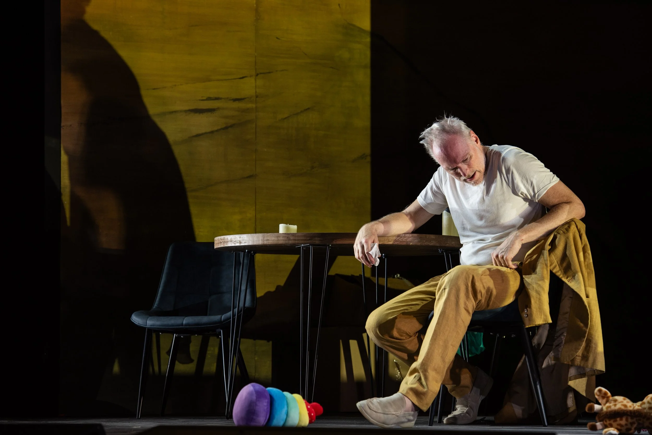 Ein Mann sitzt an einem Tisch, trägt ein weißes T-Shirt, braune Hosen und schaut nach unten. Er hält einen weißen Gegenstand in der Hand und scheint zu schlafen oder sich auszuruhen. Auf dem Boden steht eine bunte Puppe, und im Hintergrund gibt es eine große gelbe Wand. Neben dem Mann liegt eine braune Jacke, und es gibt Kerzen auf dem Tisch.