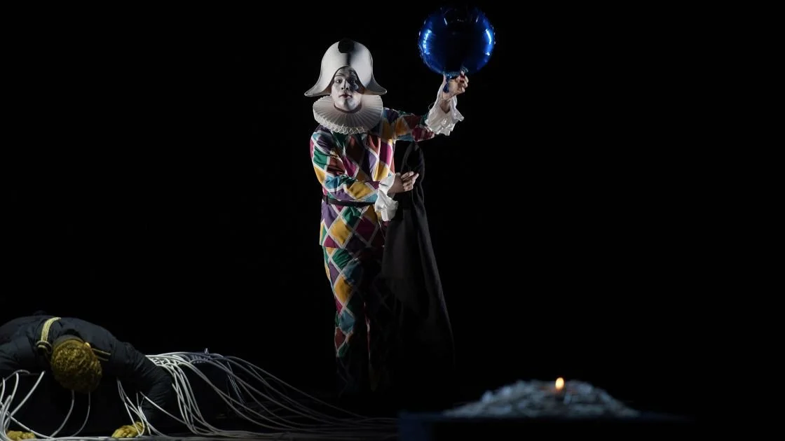 Ein Clown in bunter Kleidung, weißem Gesicht und großem weißen Hut steht auf einer dunklen Bühne. Er hält eine blaue Ballonfigur in der Hand. Vor ihm liegt eine Person, die auf dem Boden liegt und mit Kabeln verbunden ist.