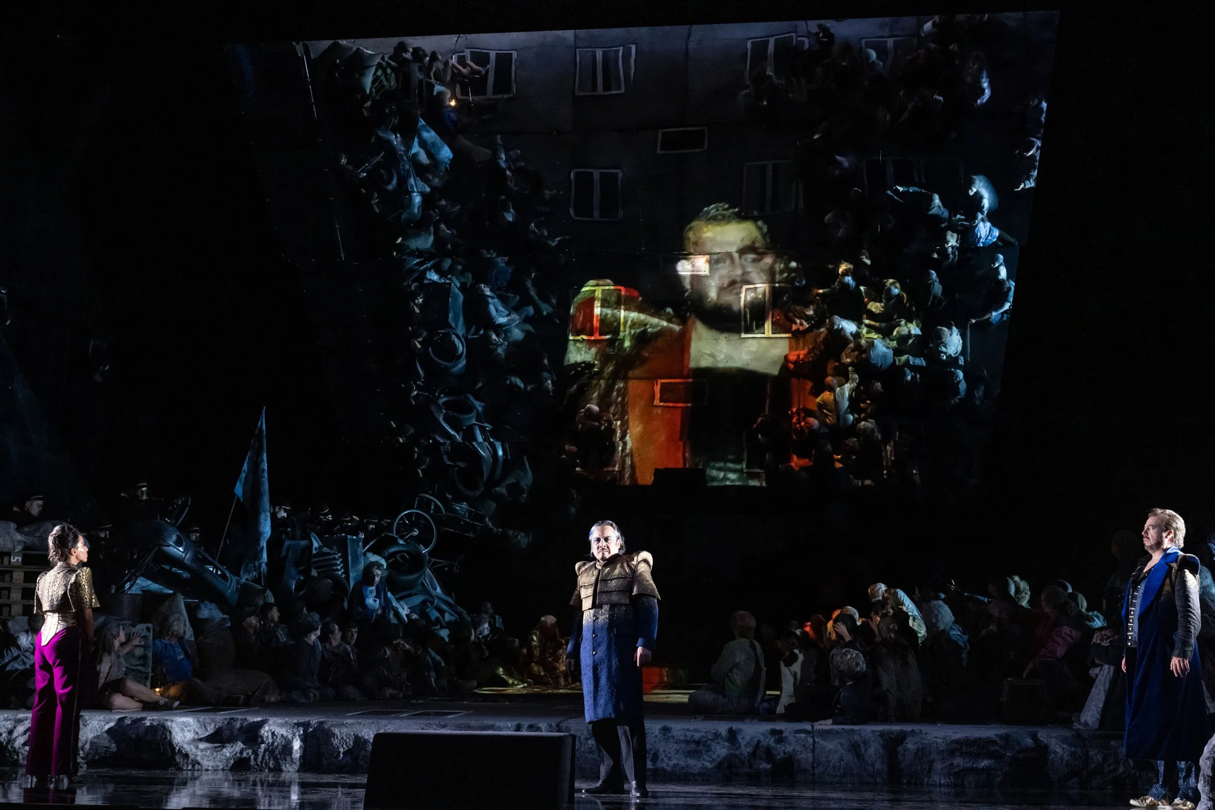 Theaterbühne mit drei Schauspielern im Vordergrund und großem Publikum im Hintergrund. Ein großer Leinwand im Hintergrund zeigt einen Mann mit Brille, vermutlich eine Projektion oder Video. Die Szene ist dramatisch, mit dunkler Beleuchtung.