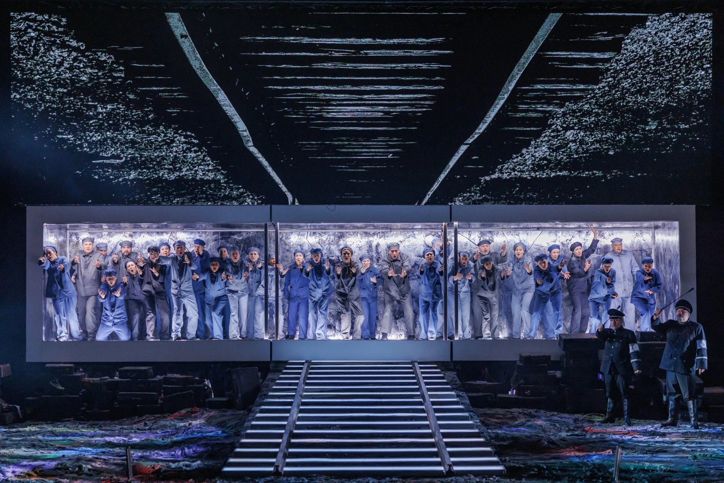 Theaterbühne mit einer Gruppe von Schauspielern in blauer Uniform, die in einem Glaskasten auftreten, im Hintergrund eine große Projektionsfläche mit abstrakten weißen Linien auf schwarzem Hintergrund.