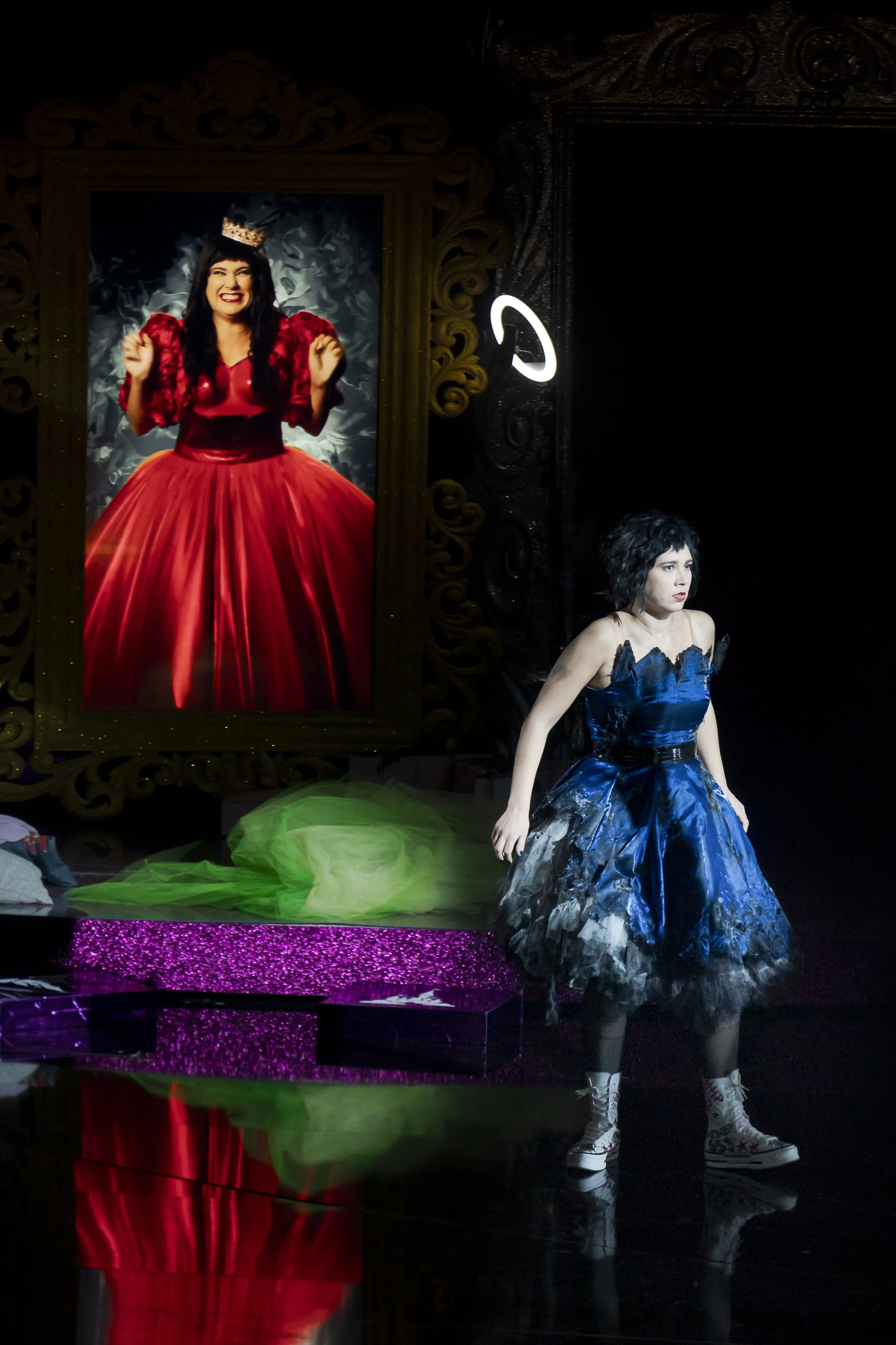 Eine Frau in einem blauen Kleid auf einer Bühne mit einem großen Bild von einer Frau in einem roten Kleid und Krone im Hintergrund.