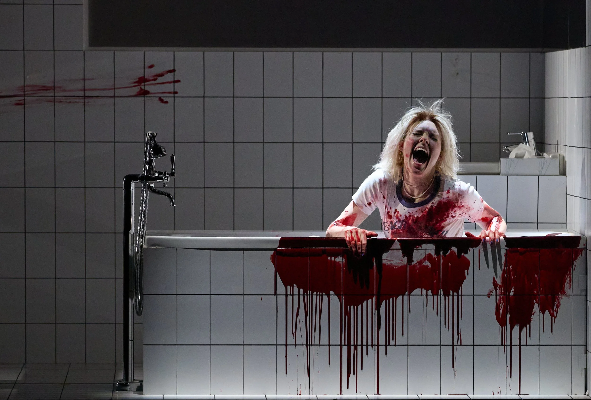 Eine erschrockene Frau mit blonden Haaren in einer blutverschmierten weißen T-Shirt sitzt in einer Badewanne, die mit Blut bedeckt ist, in einer dunkel beleuchteten Badezimmerumgebung.