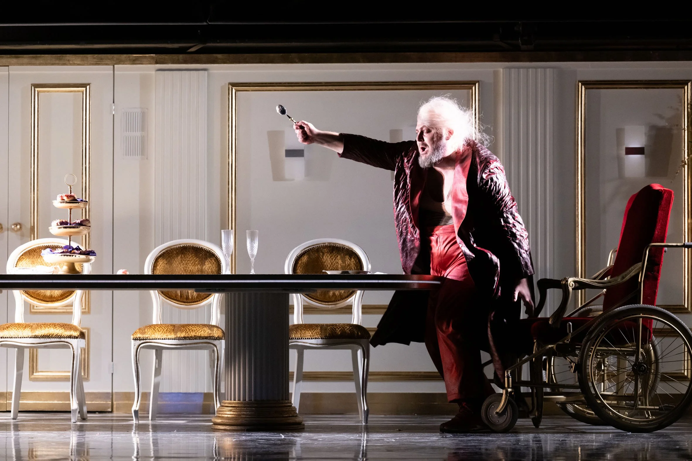 Ein älterer Mann mit weißem Haar und Bart, in einem Theaterkostüm, steht vor einem Esstisch und streckt einen Löffel aus, während er an einem Rollstuhl sitzt, in einem elegant möblierten Raum.