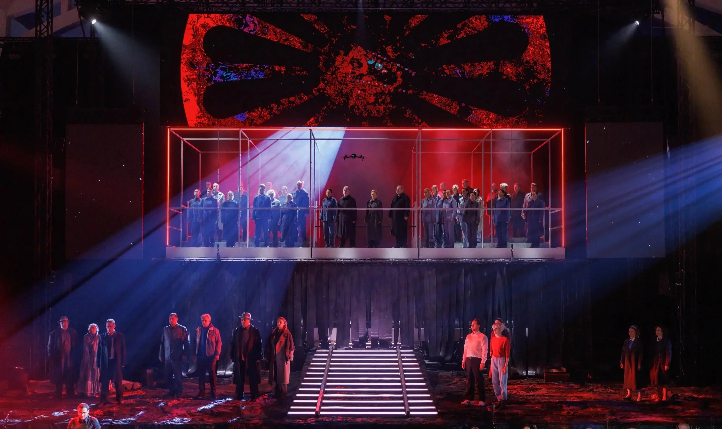 Bühne mit mehreren Menschen in zwei Ebenen, neonbeleuchteter Rahmen, dramatische Beleuchtung, rotes und blaues Licht, große LED-Leinwand im Hintergrund mit rotem und schwarzem Design.