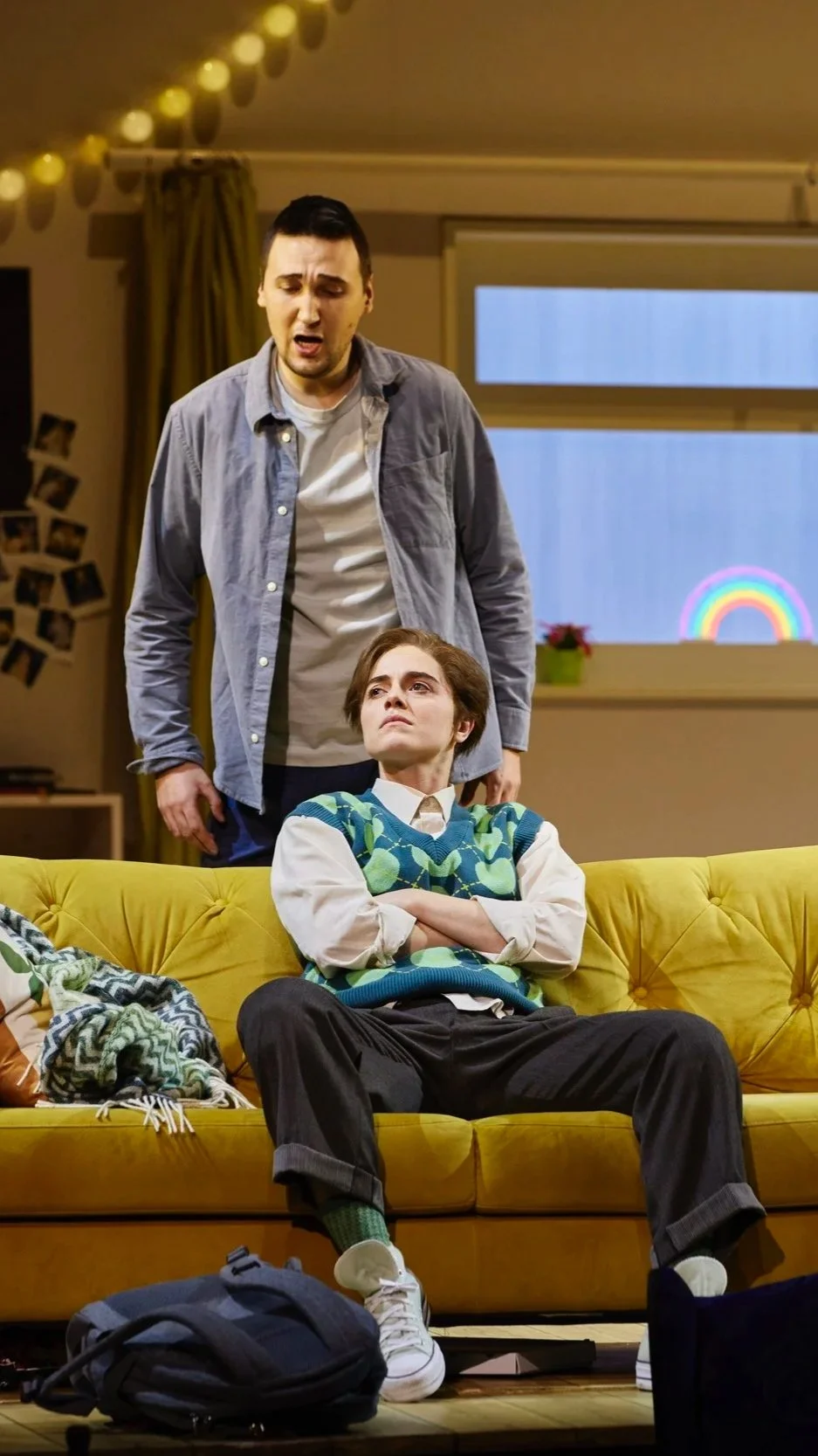 Zwei Männer, einer sitzend auf einer gelben Couch mit verschränkten Armen und skeptischem Blick, der andere steht hinter ihm und spricht, im Wohnzimmer mit Lichtergirlande und Fensterscheibe, Regen im Hintergrund.