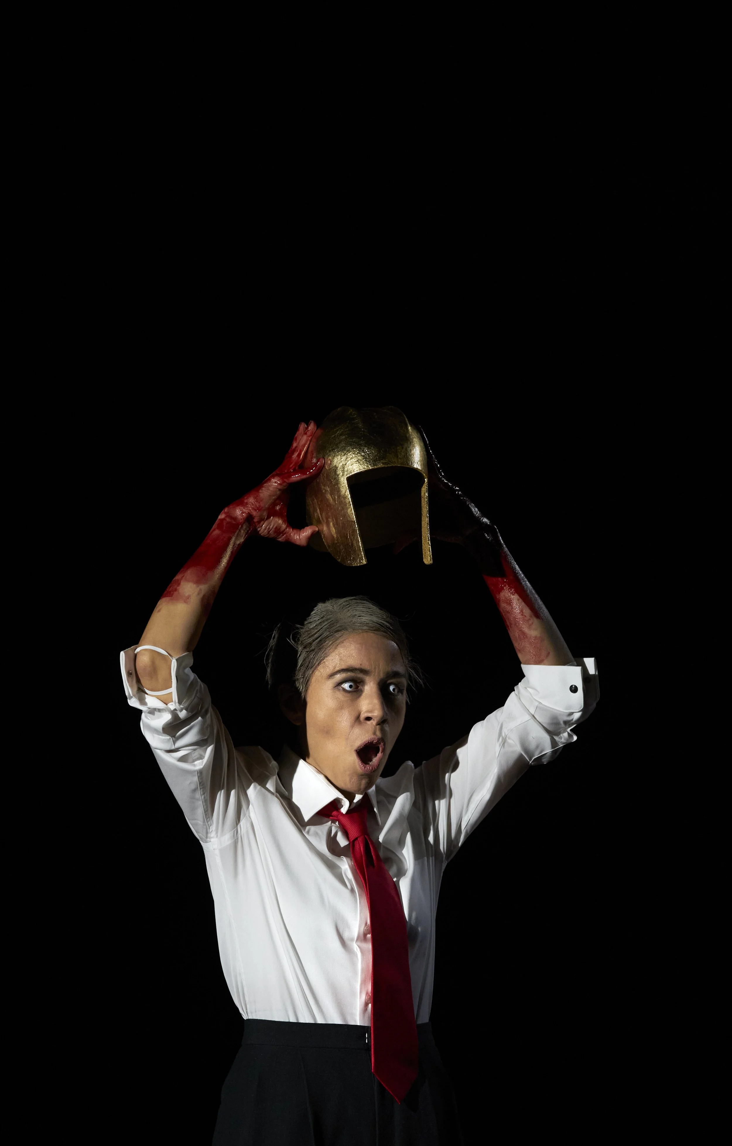 Eine Frau in Schuluniform mit roter Krawatte hält eine goldene Ritterrüstung in der Hand und sieht überrascht oder schockiert aus, vor schwarzem Hintergrund.