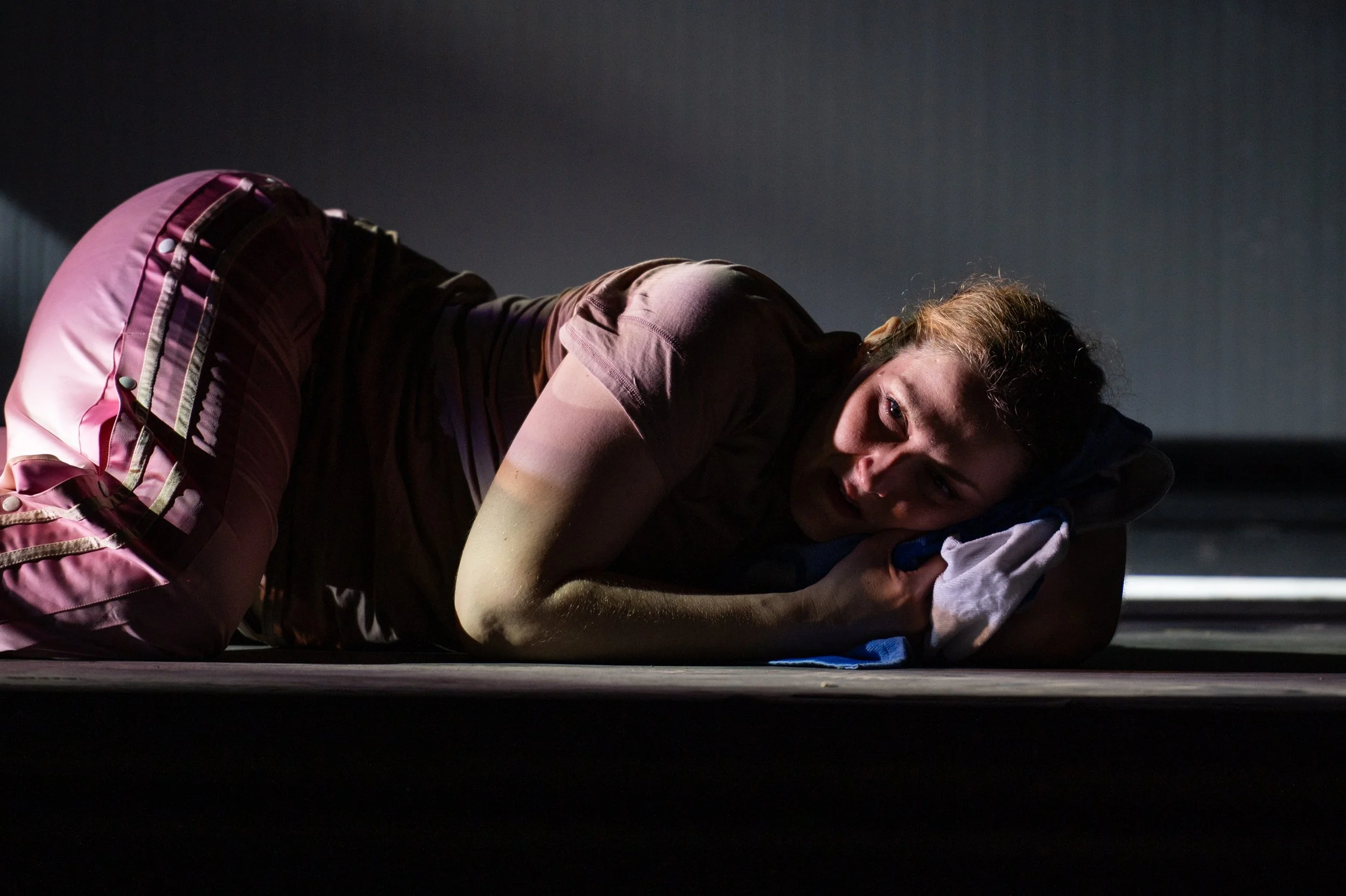 Frau liegt auf dem Boden, hält ein Handtuch als Kissen und schaut ängstlich. Dunkler Hintergrund, dramatische Beleuchtung.