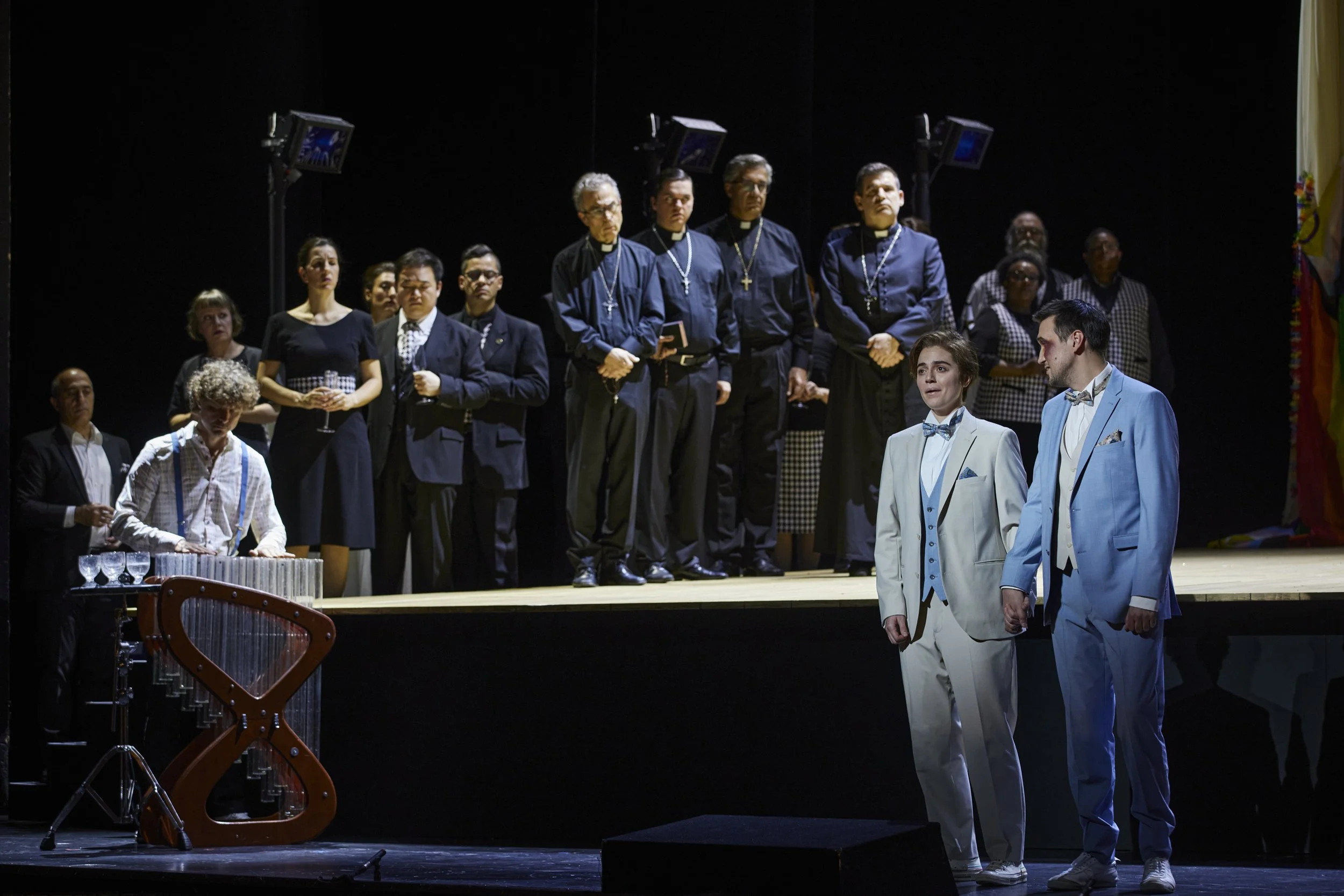 Schauspieler auf einer Theaterbühne während einer Aufführung, einige stehen in formeller Kleidung, andere im Hintergrund in dunklen Outfits. Zwei Schauspieler in weißen Anzügen stehen vorne, einer mit braunen, der andere mit schwarzen Haaren, während eine Person auf der linken Seite Klanginstrumente spielt.