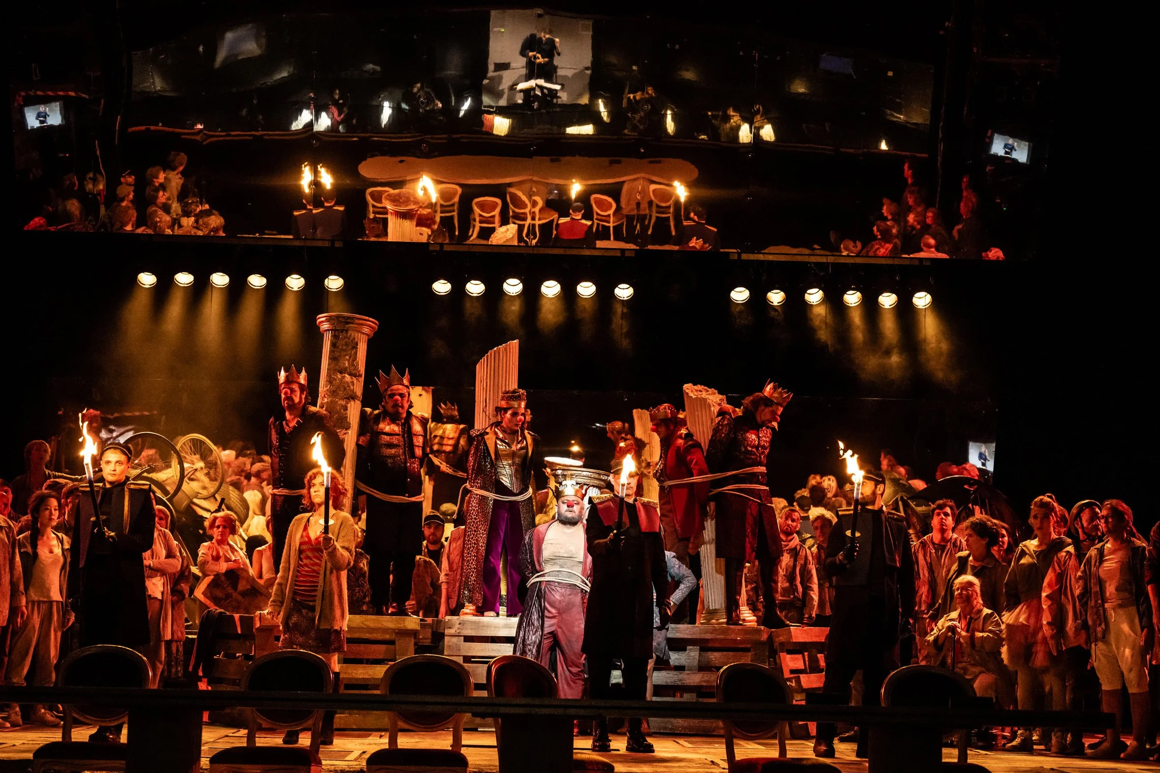 Bühnenbild eines Theaters mit Menschen in Kostümen, inklusive Königs- und Erobererkostümen, auf einer improvisierten Treppe, mit Feuer- und Kerzenlicht, bei einer Inszenierung oder Performance.
