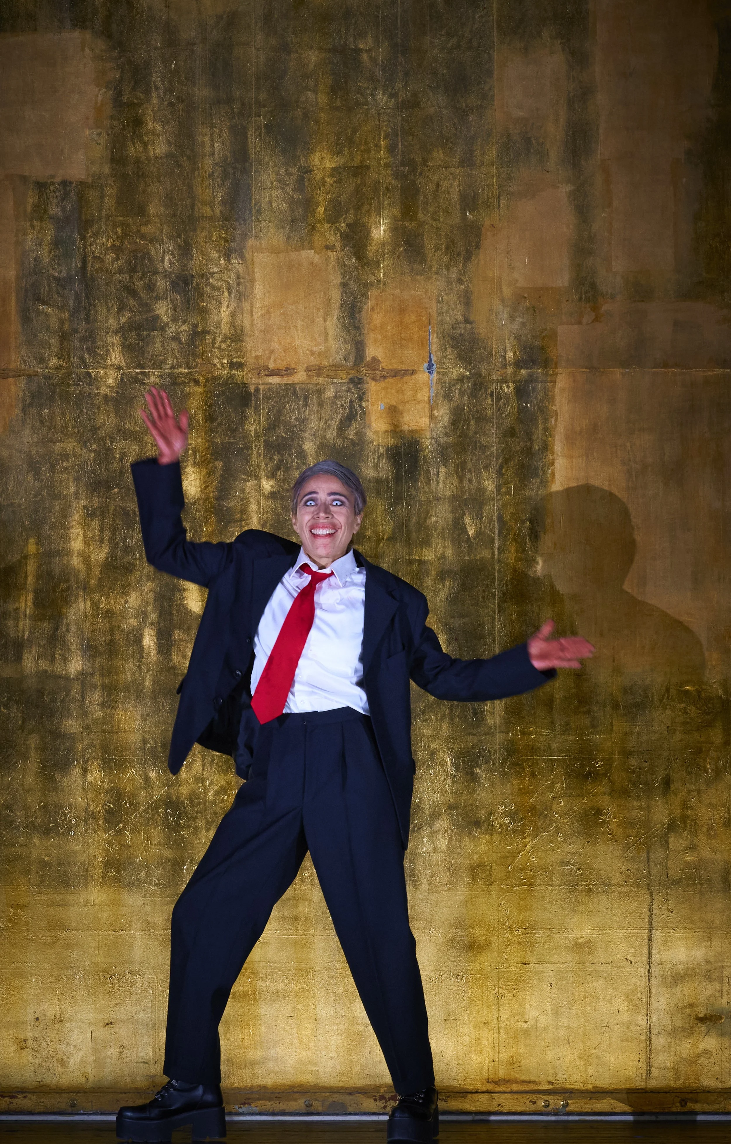 Menschen auf einer Bühne, die in ein Mikrofon spricht und eine dramatische Haltung einnimmt, vor einer goldfarbenen Hintergrundwand.