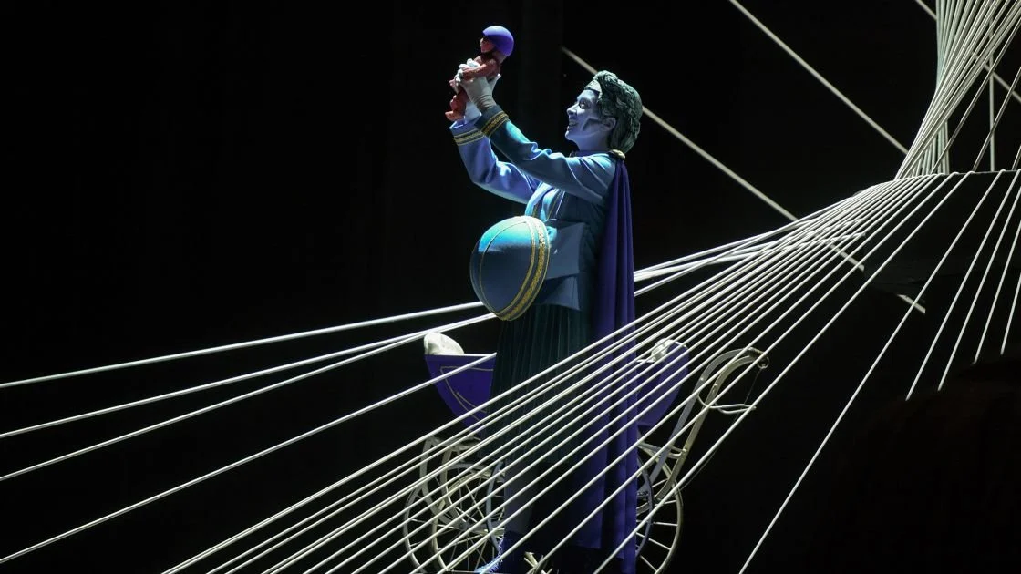 Eine Figur, die einen Mann darstellt, der auf einem Fahrrad sitzt und ein Baby hochhält, um es zu küssen, mit einem schwarzen Hintergrund und weißen Seilen im Vordergrund.