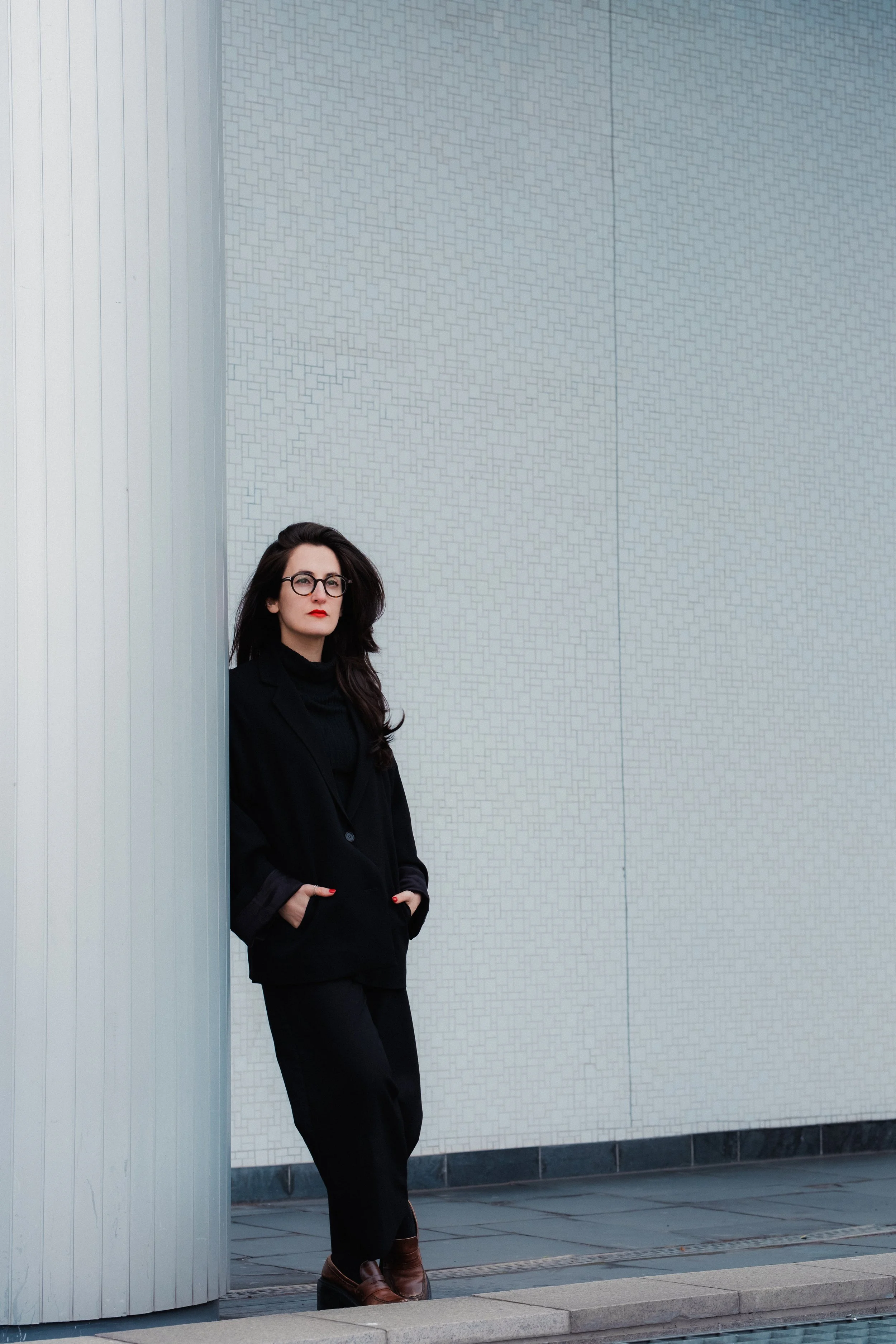 Frau mit schwarzen Haaren, Brille, rotem Lippenstift und dunklem Anzug steht vor einer modernen Wand aus weißen Fliesen und einem Metallrohr, außerhalb auf einer gepflasterten Fläche.