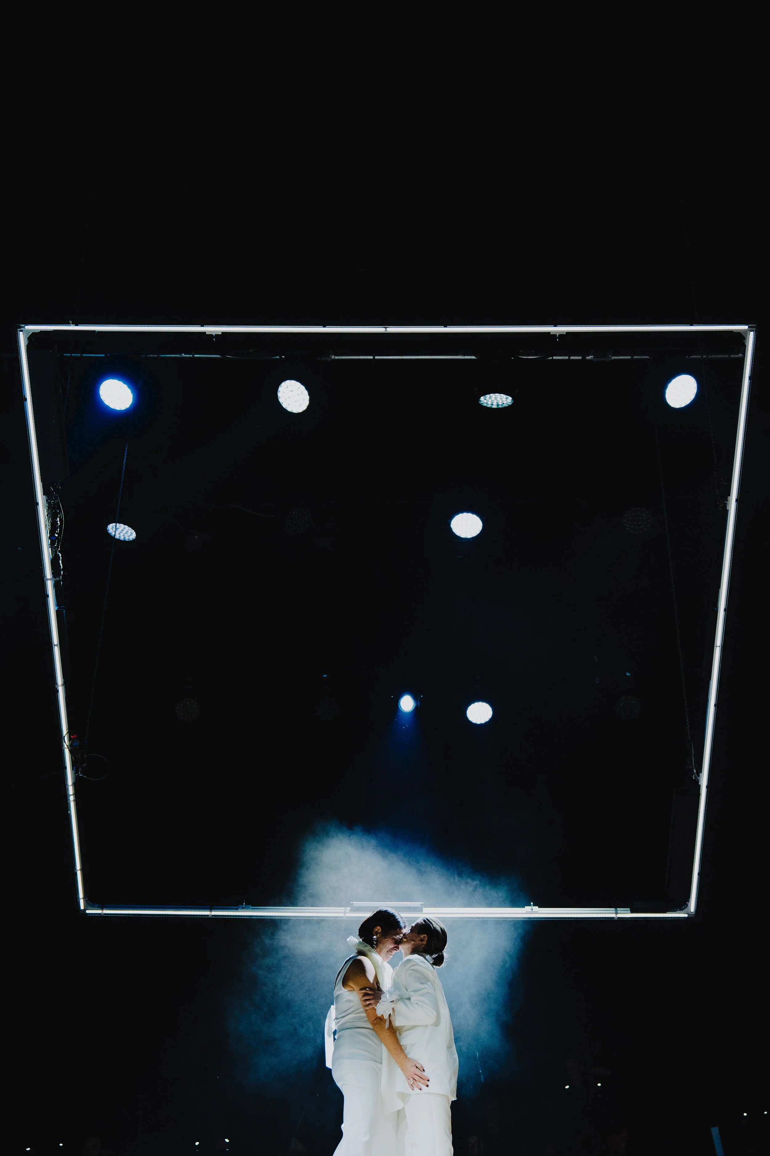 Zwei Frauen in weißen Outfits stehen einander gegenüber unter einem dreieckigen Rahmen mit blauen und weißen Lichtern, im Dunkeln, bei einer Beleuchtungsshow.