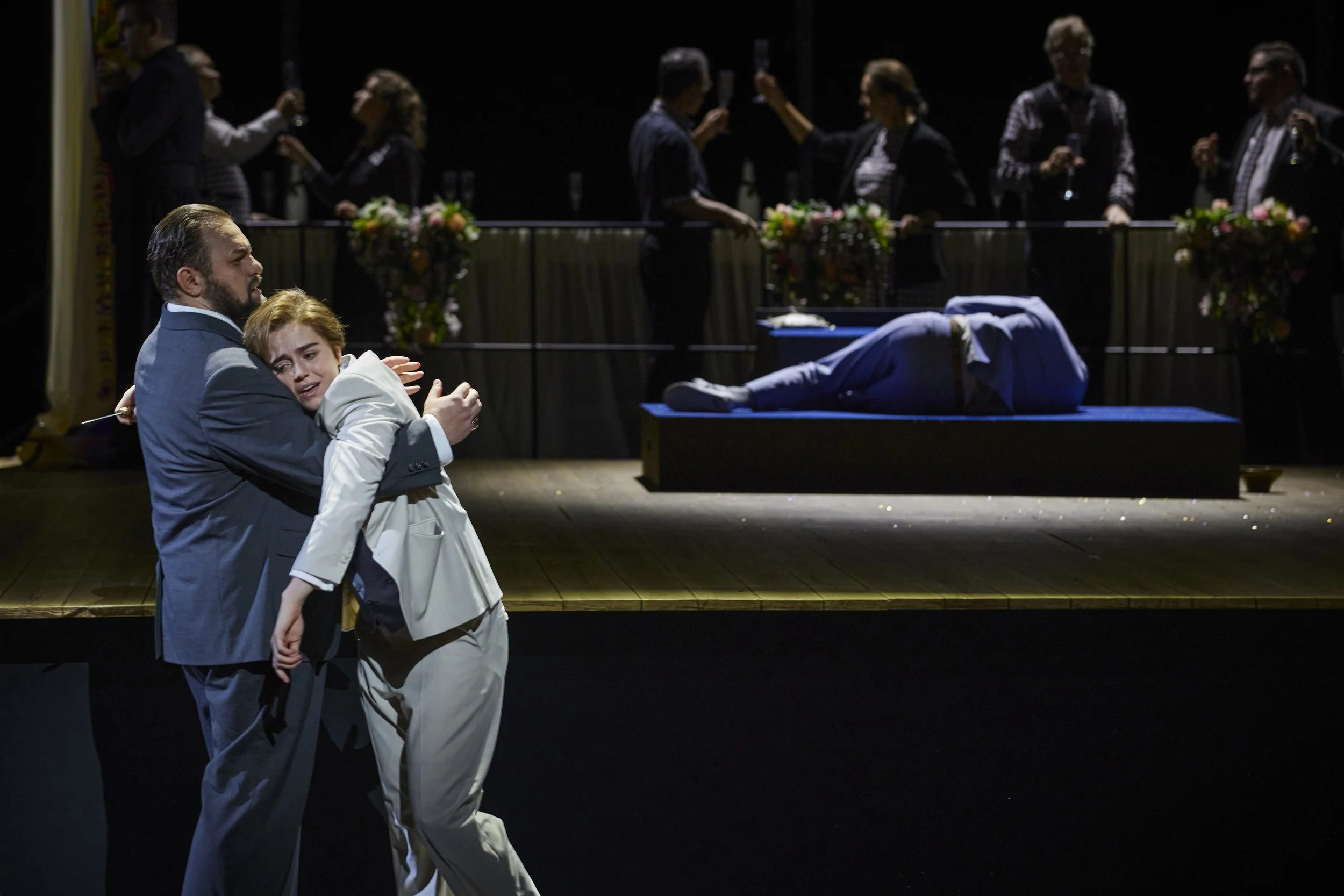 Eine dramatische Bühnenszene, in der zwei Menschen im Vordergrund emotional miteinander interagieren, während im Hintergrund eine Leiche auf einer Bühne liegt. Es gibt mehrere Personen, die auf einer erhöhten Plattform in der Nähe einer Bar anstößige Getränke trinken, und farbenfrohe Blumendekorationen.