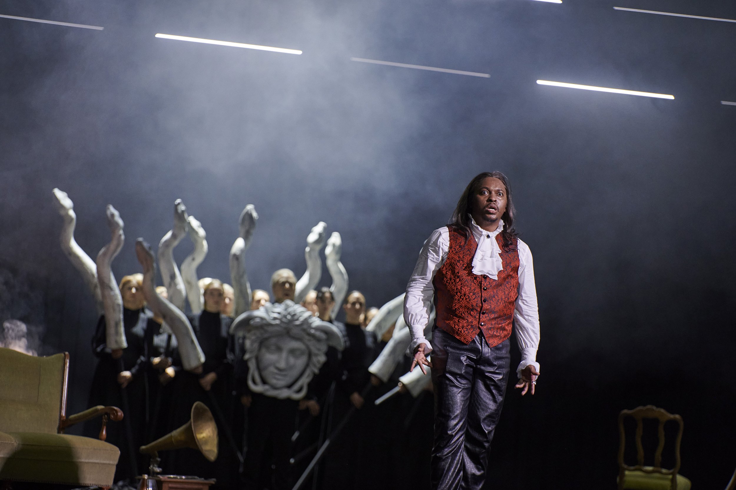 Ein Mann in viktorianischer Kleidung steht auf einer Bühne, im Hintergrund eine Gruppe von Menschen in schwarzen Anzügen mit weißen Schals und mehreren weißen ausgestreckten Armen. Es gibt alte Möbelstücke und eine schwarze Statue auf der Bühne, und 