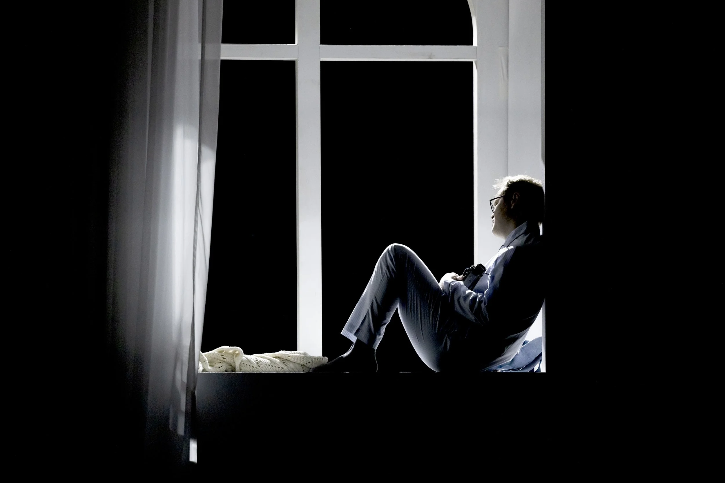 Junge Frau sitzt am Fenster, blickt nach draußen, mit Brille und Kamera in der Hand, im Schatten, Blick nach rechts, nächtliche Szene.