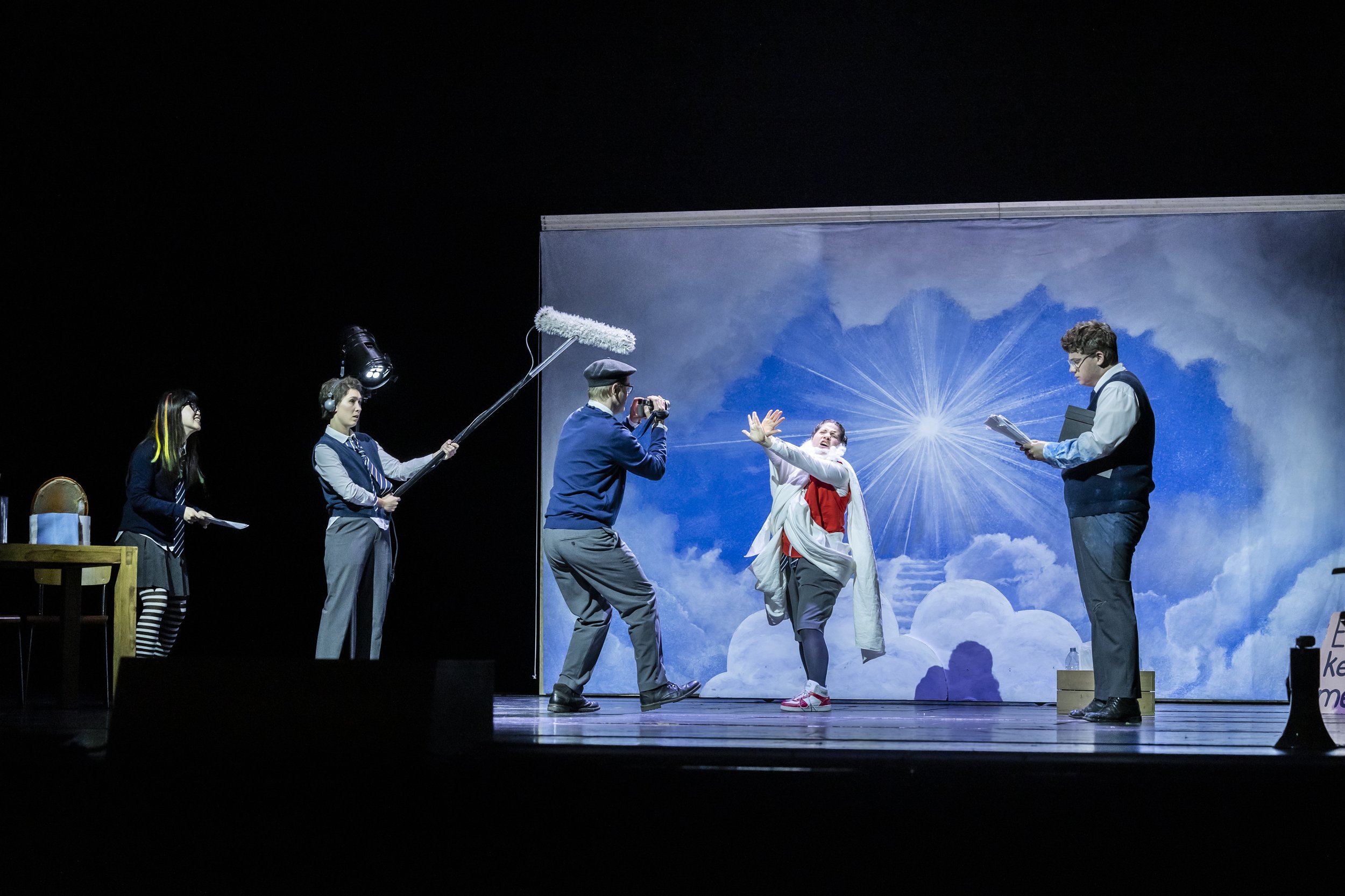 Bühnenszene aus einem Theaterstück, mit fünf Schauspielern, einer mit Bohrmaschine, eine mit Wischer, eine fotografiert, eine liest ein Skript, eine macht Gesten, mit einer Himmelshintergrundprojektion.