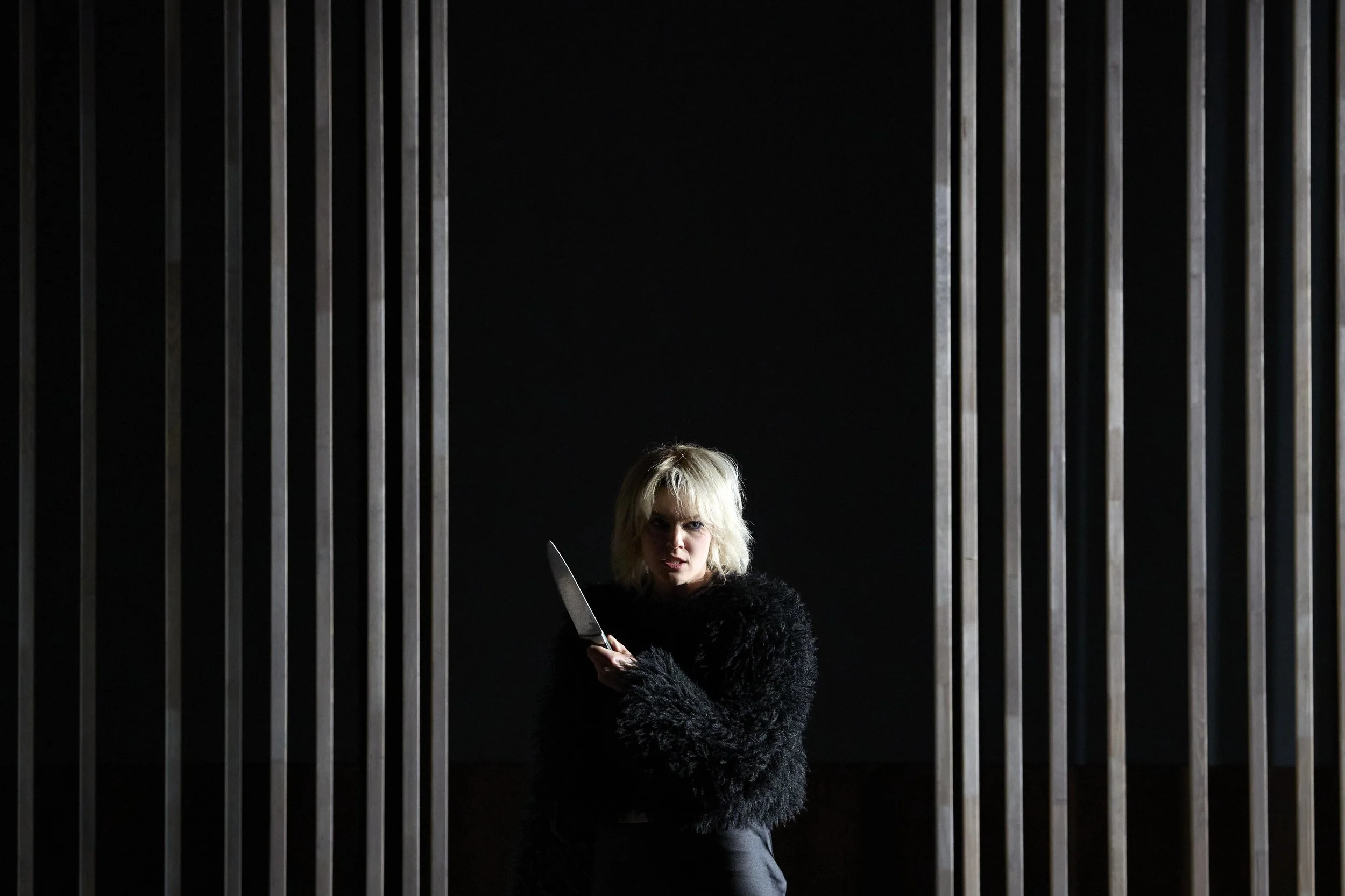 Frau mit blonder Haart, schwarze Jacke, hält ein Messer, steht vor vertikalen Metallstäben in einem dunklen Raum.
