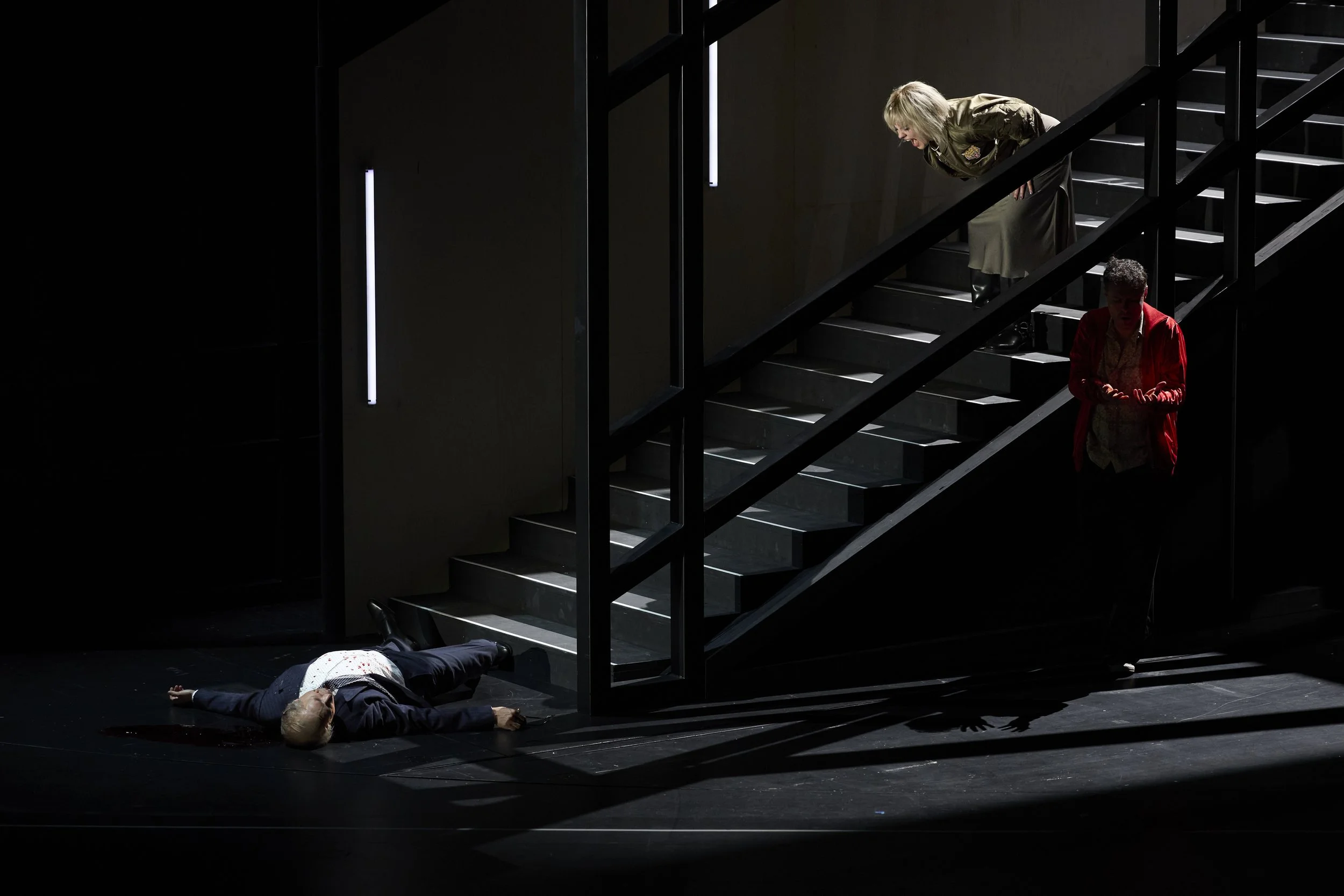 Drei Menschen auf einer dunklen Bühne: Eine Person liegt auf dem Boden, während die anderen beiden Personen auf der Treppe stehen und zu ihr schauen.