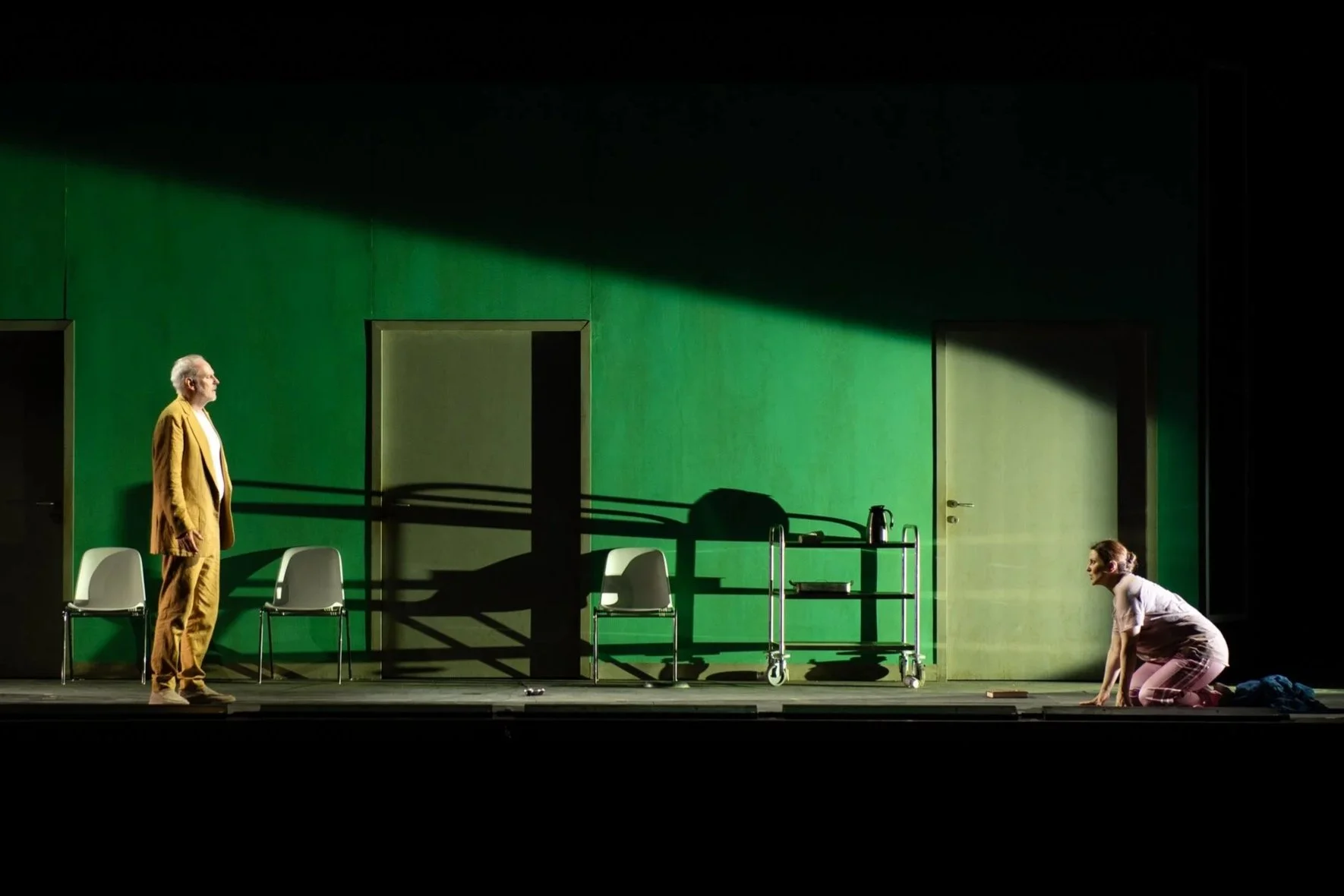 Zwei Menschen auf einer Bühne, eine Frau kniet auf dem Boden, ein Mann steht gegenüber, vor einer grünen Wand.