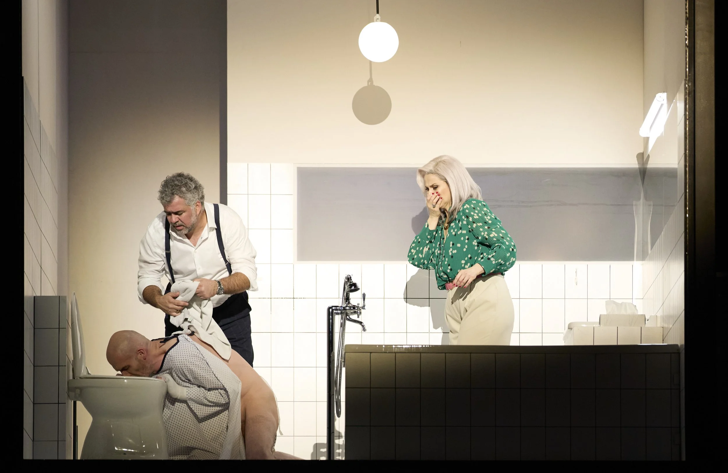 Drei Menschen in einem weißen Badezimmer, einer sitzt auf der Toilette, der zweite steht neben ihm und hält ein Handtuch, die dritte Frau steht am Wannenrand und lacht.