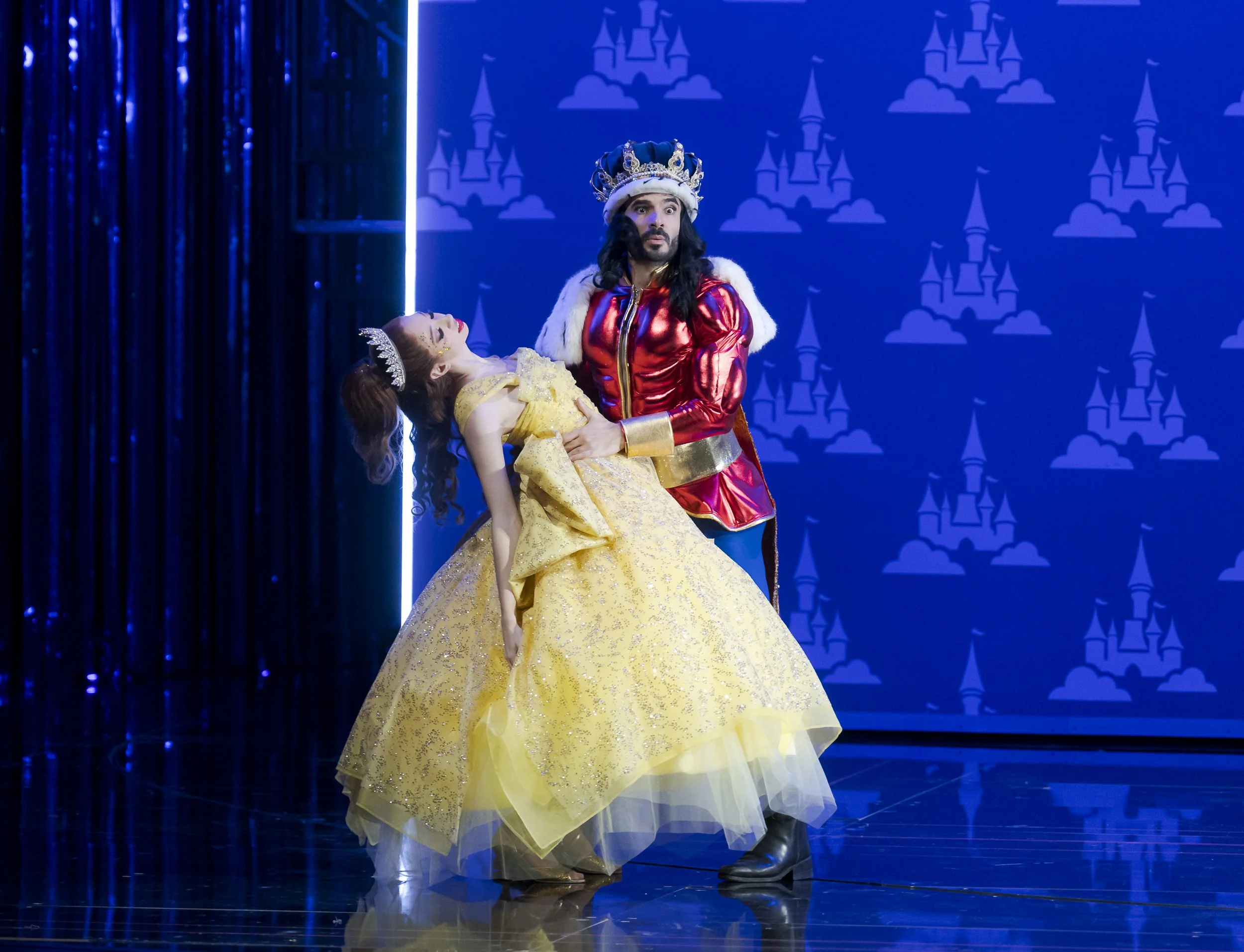 Ein Bühnenbild mit zwei Menschen in Kostümen. Eine Frau in einem goldgelben Kleid und eine Krone liegt im Arm eines Mannes im rotem Kronen- und Schulter-Kostüm. Im Hintergrund eine blaue Wand mit Schloss-Design.