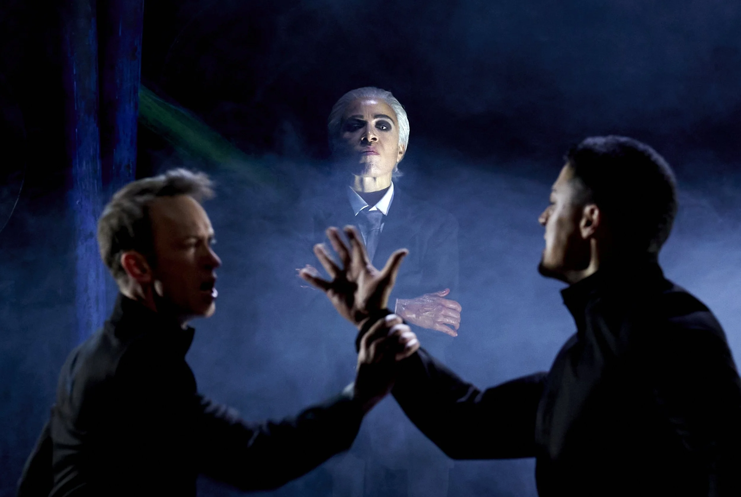 Drei Männer in dunkler Kleidung, die sich gegenüberstehen und sich die Hände schütteln, im Hintergrund eine schattenhafte Figur mit weißem Haar, die in einen dunklen Raum blickt.