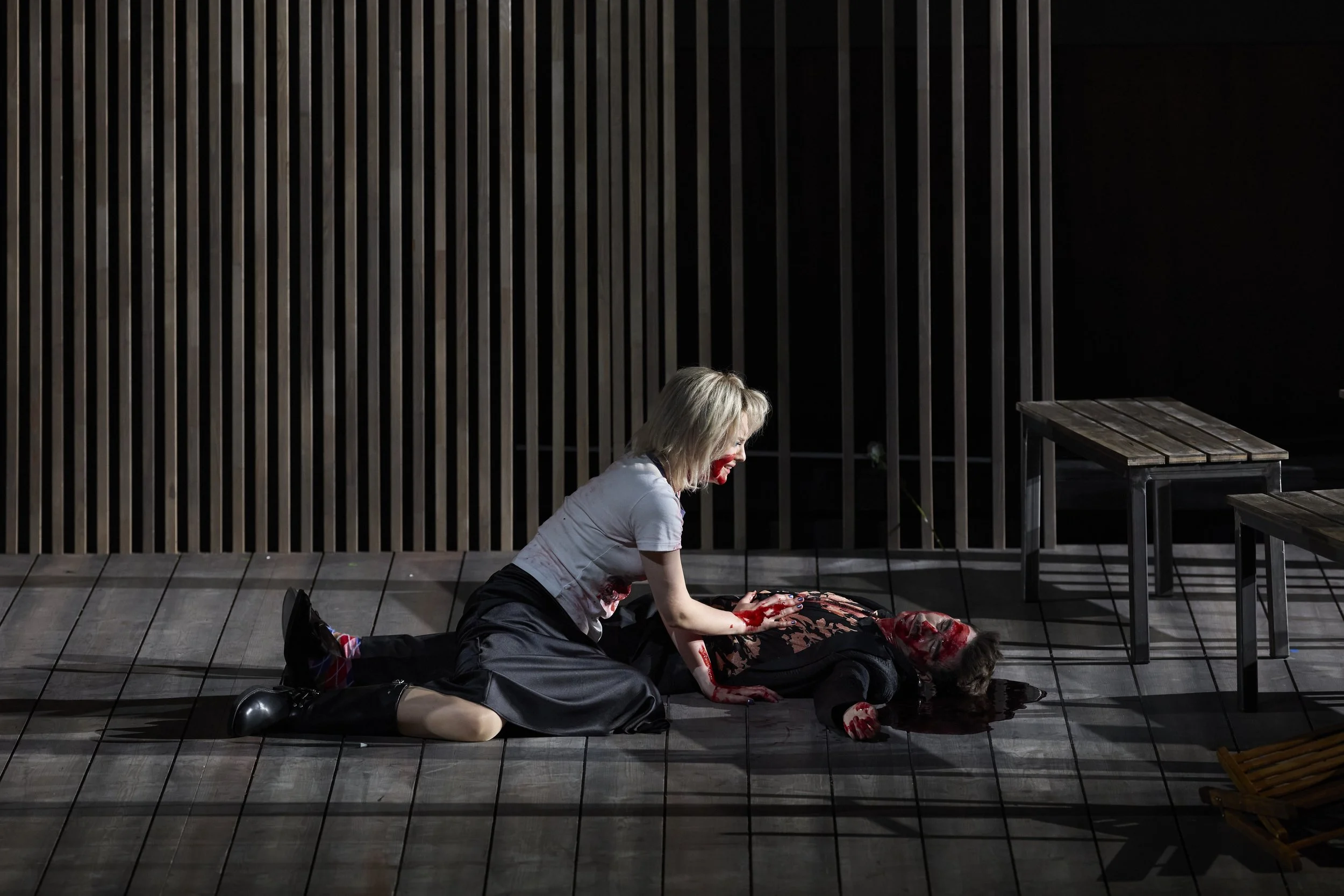 Bühnenaufnahme eines Horrorspiels, bei dem eine Schauspielerin auf dem Boden liegt, sehr blutig aussieht und von einer anderen Person tröstend berührt wird, vor einem dunklen, modernen Bühnenhintergrund.