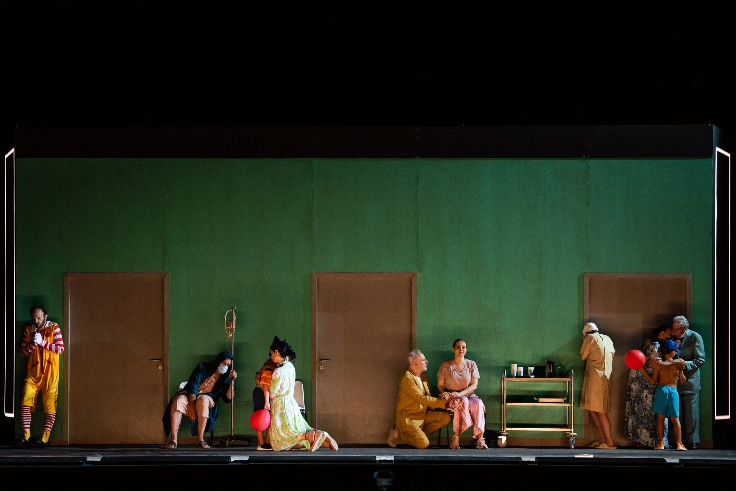 Bühnenbild einer Theaterproduktion mit mehreren Schauspielern in verschiedenen Szenen, einige sitzen, andere stehen, vor grüner Wand mit drei Türen, einige sind in farbenfrohen Kostümen, eine Person trägt eine medizinische Maske, eine hält einen rote