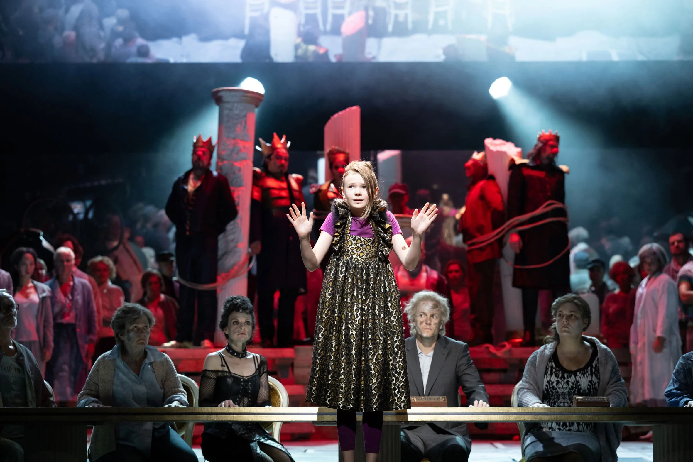 Junge Schauspielerin steht auf einer Bühne in einem Theaterstück, umgeben von anderen Schauspielern und Publikum. Im Hintergrund sind dramatische Bühnenelemente und Kostüme zu sehen.