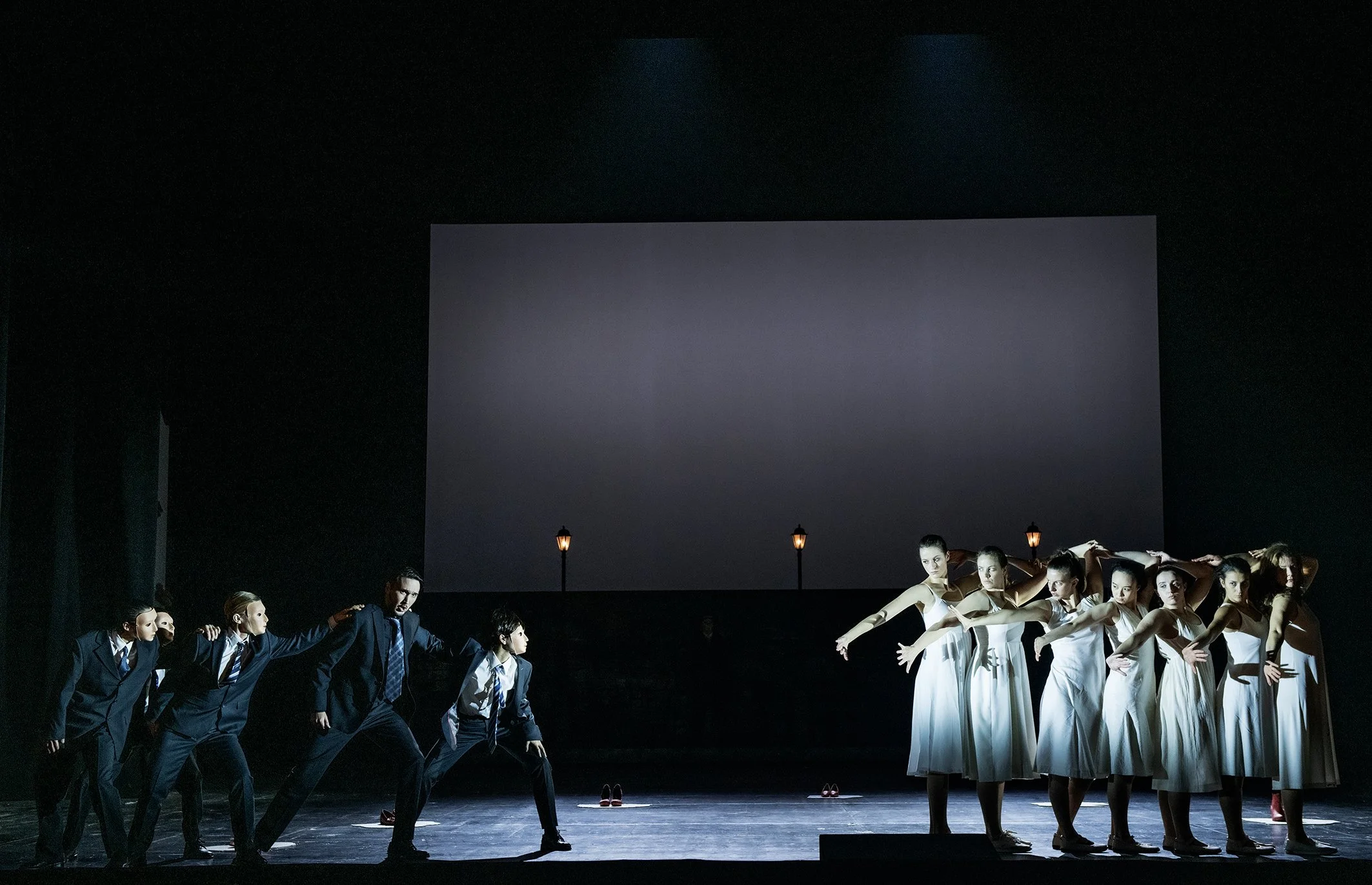 Balletttänzer in weißen Kleidern auf der Bühne, gegen einen dunklen Hintergrund mit einer großen leinwandartigen Fläche und Straßenlaternen