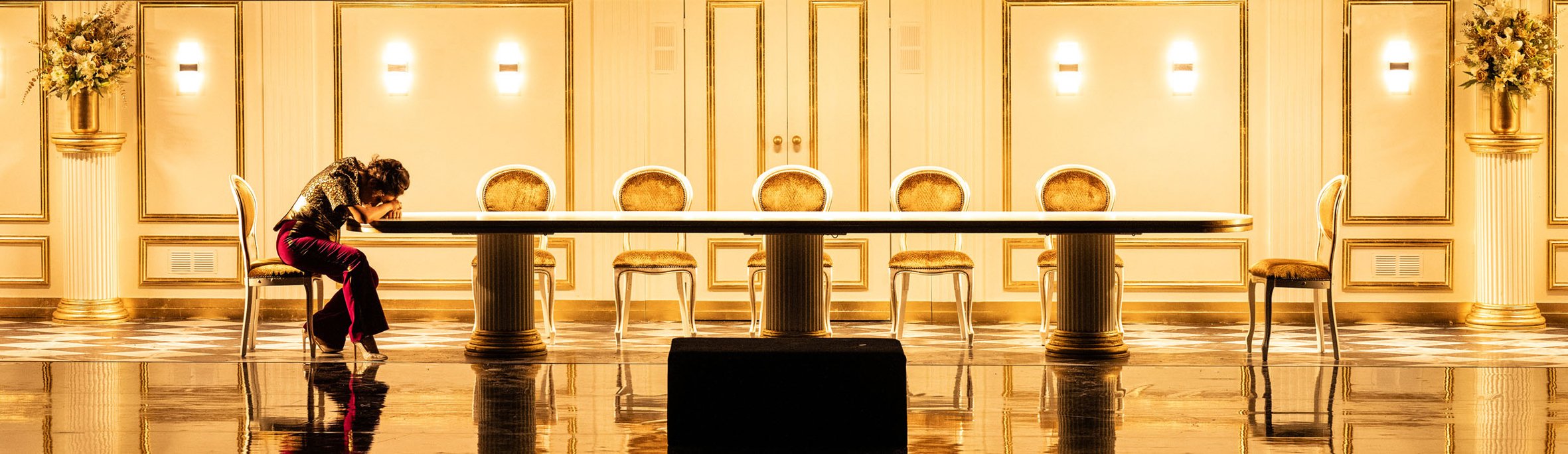 Leere goldene und weiße elegante Festtafel mit sieben Stühlen in einem prunkvollen Raum, links eine Frau mit langen dunklen Haaren, grauem Oberteil, roten Hosen und schwarzen Schuhen sitzt mit vorgeneigtem Kopf an einem Stuhl.