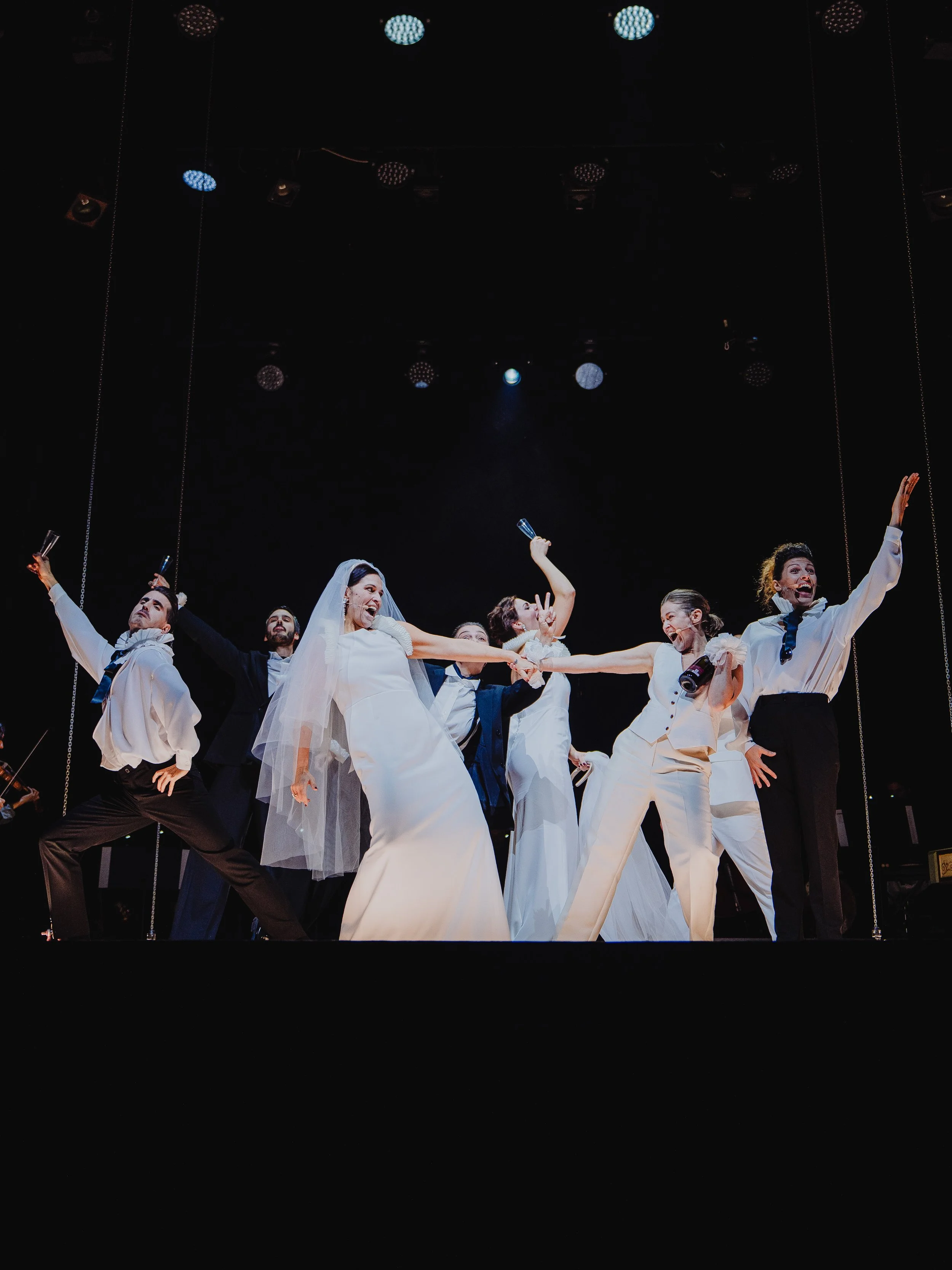 Schauspieler auf Bühne, die als Braut und Bräutigam verkleidet sind, umgeben von anderen Schauspielern in Gespräche und Posen.