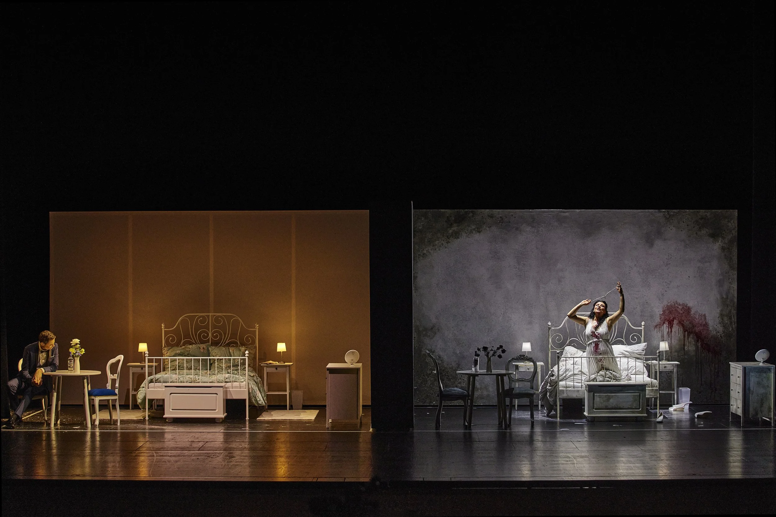 Bühnenaufnahme eines Theaterspiels mit zwei Szenen, links ein Mann an einem Tisch, rechts eine Frau im weißen Kleid im Schlafzimmer mit grauem Hintergrund